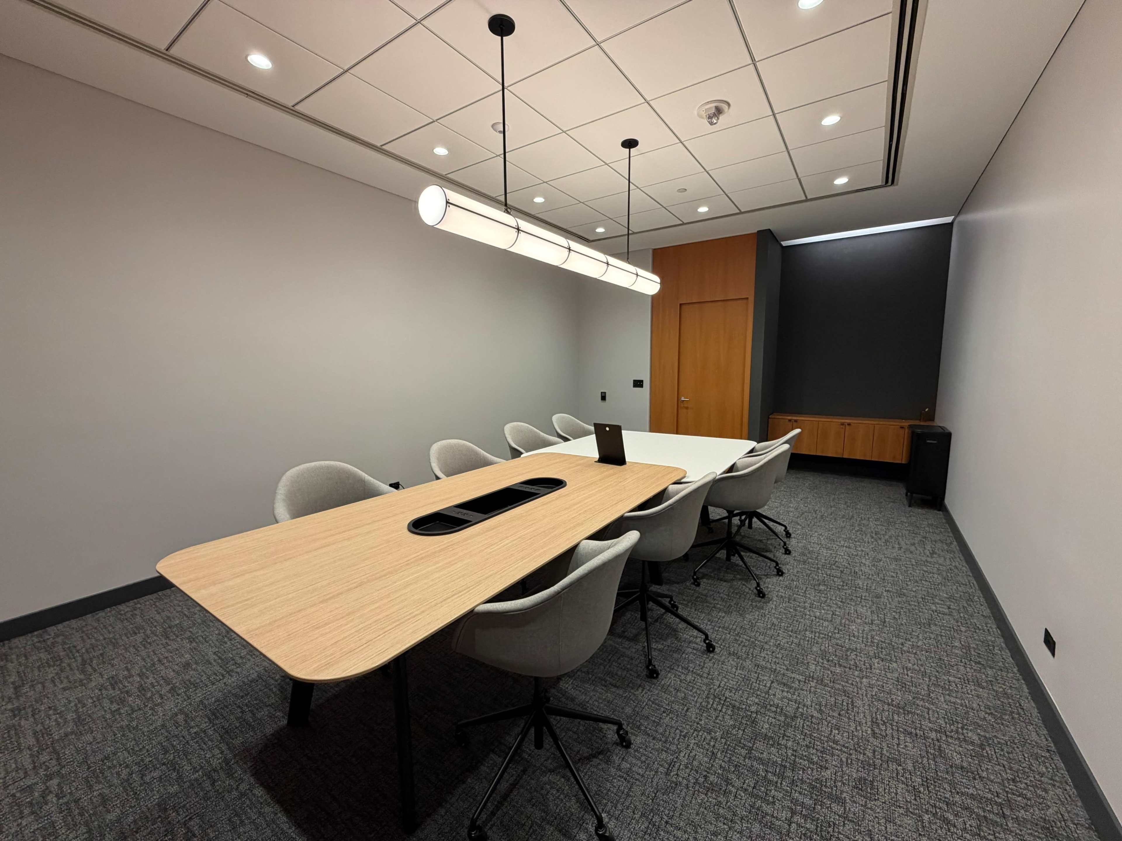 A modern conference room features a long wooden table surrounded by six ergonomic chairs, with a large light fixture above and a wall-mounted cabinet in the background.