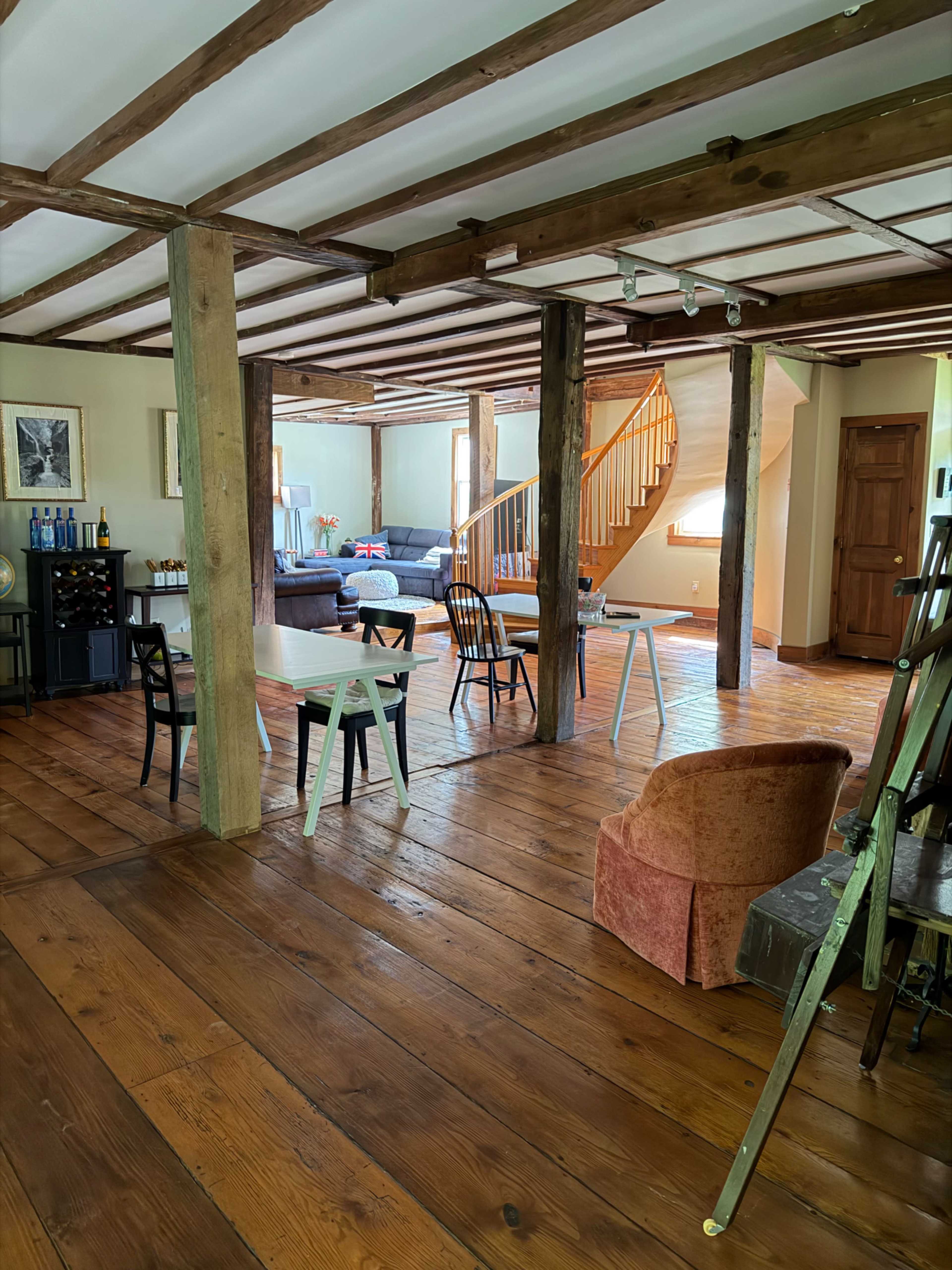 The image shows an open living space with wooden beams, a wooden floor, a spiral staircase, and various seating arrangements, including chairs and a sofa.