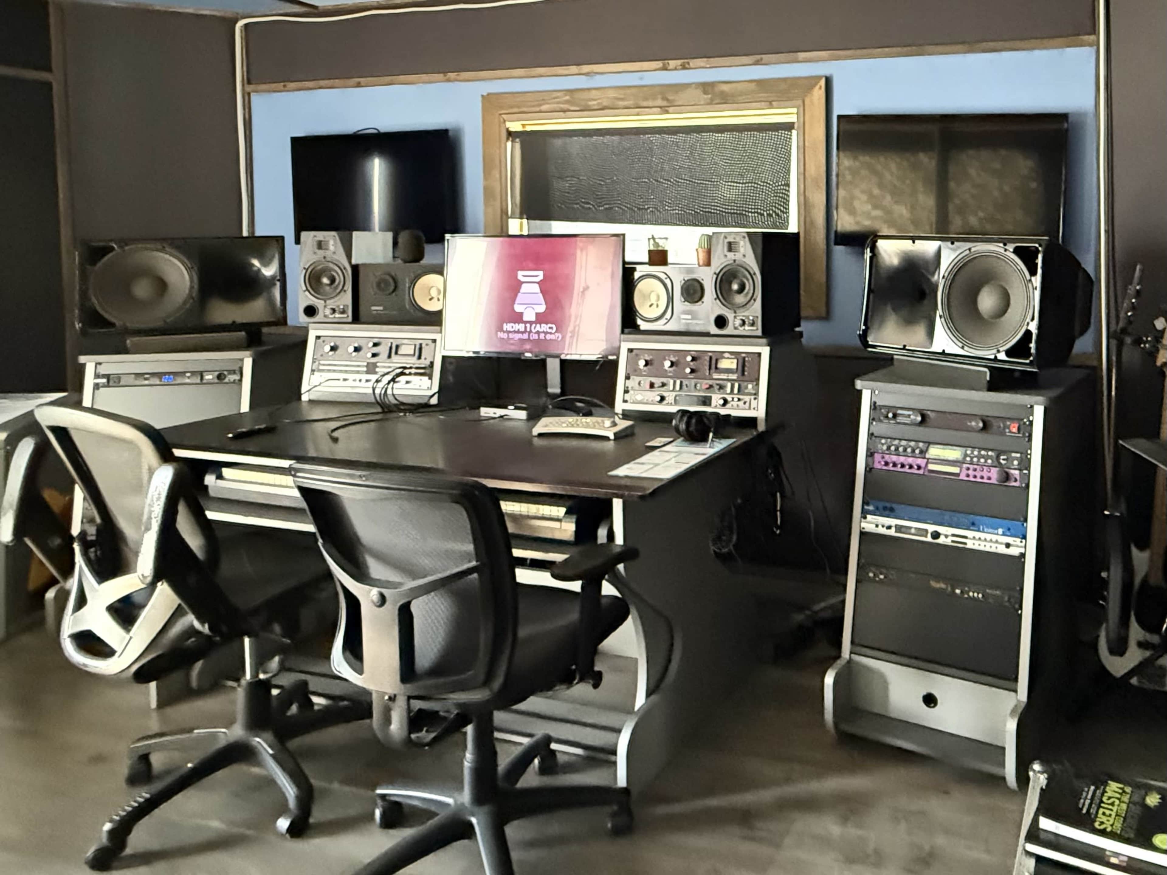 The image shows a recording studio featuring a central desk with two office chairs, audio equipment, and speakers on either side, along with a computer monitor displaying audio software.
