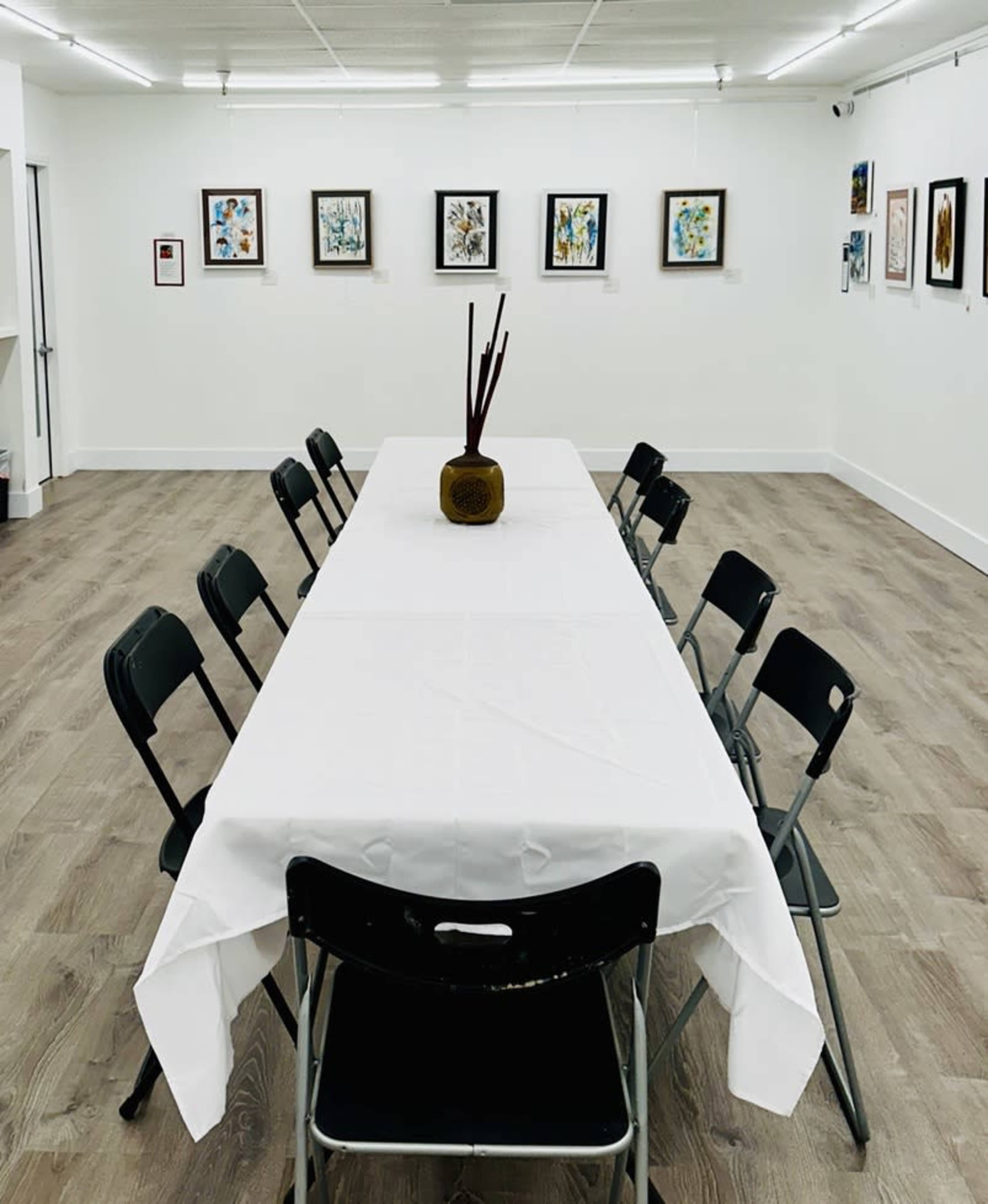 A long table with a white tablecloth is set in a bright room with artwork hanging on the walls and black folding chairs arranged around it.