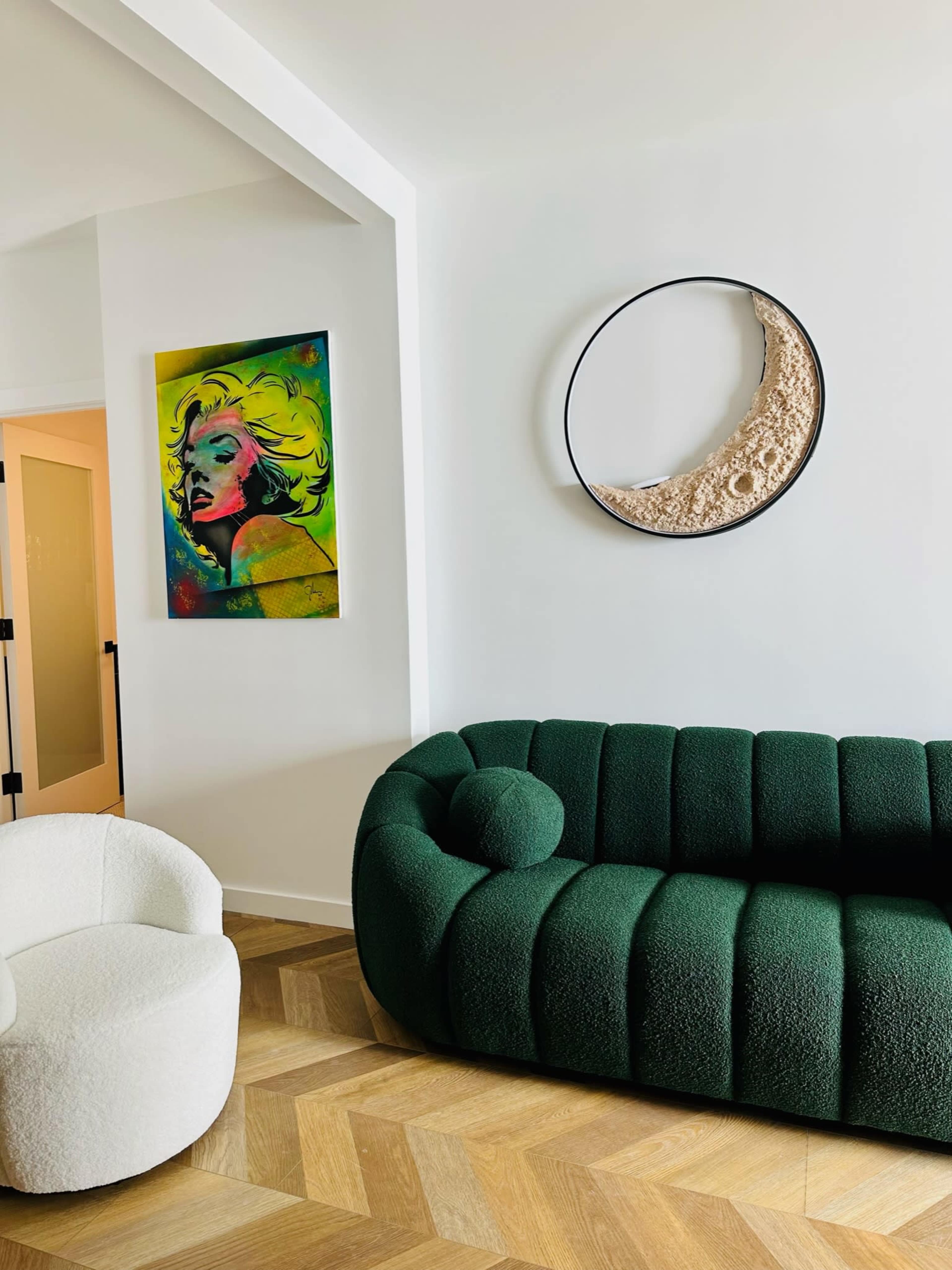 A modern living area featuring a green rounded sofa, a white armchair, a colorful artwork of a woman on the wall, and a circular decorative mirror.