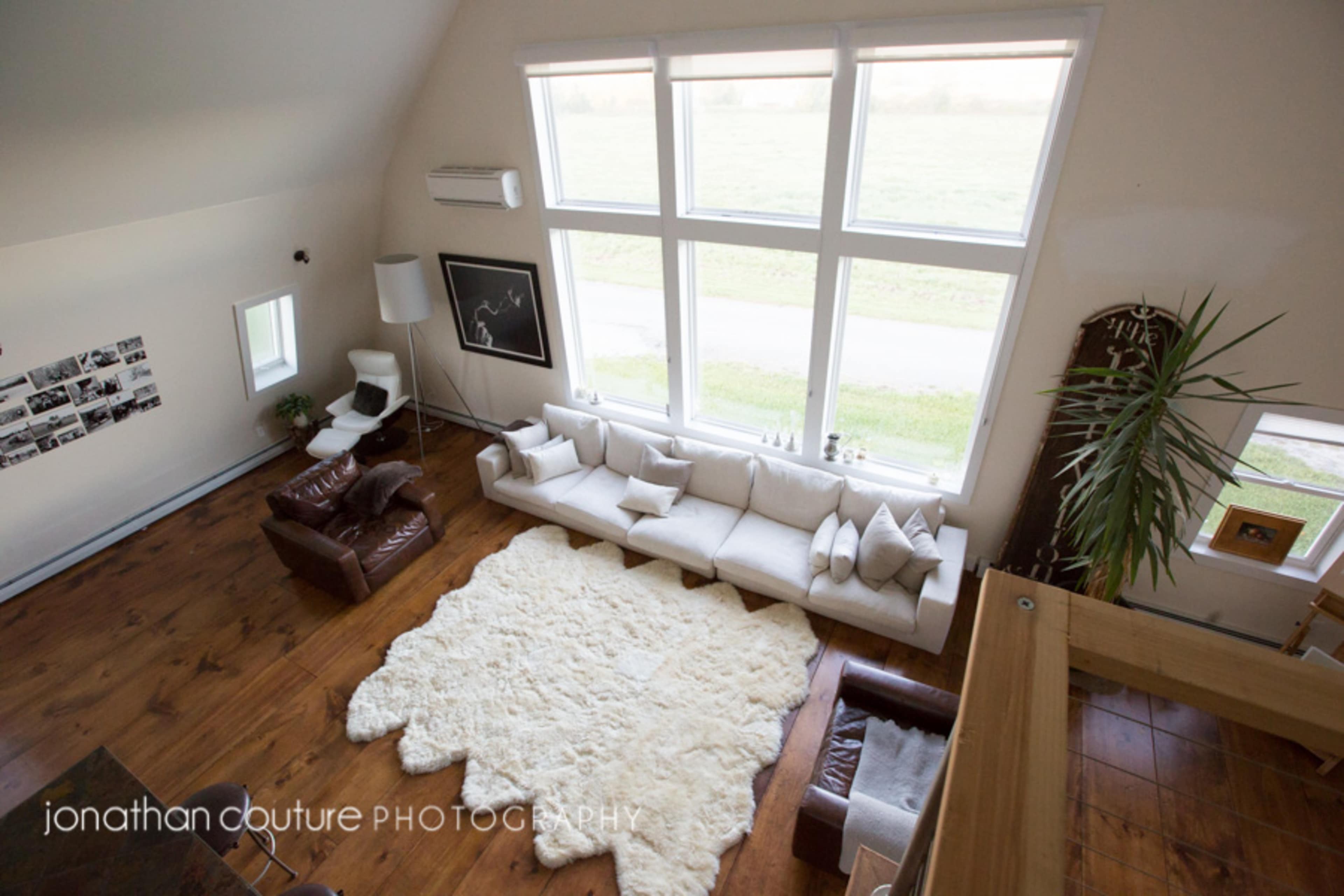 A spacious living room features large windows, a cream-colored sofa, a brown leather chair, and a textured rug on a wooden floor.