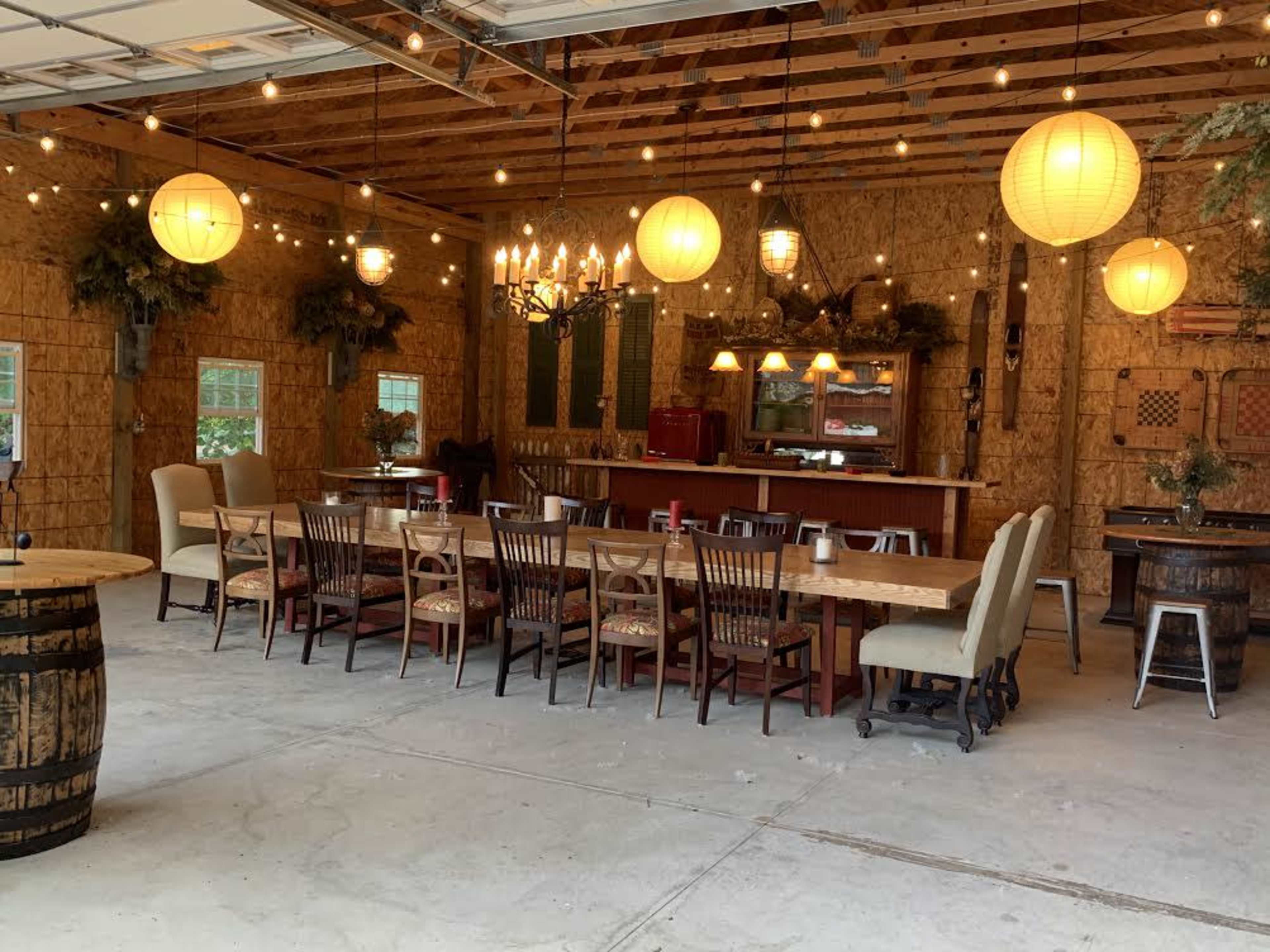 A spacious interior features a long dining table surrounded by chairs, with hanging lanterns and a chandelier illuminating the rustic decor.