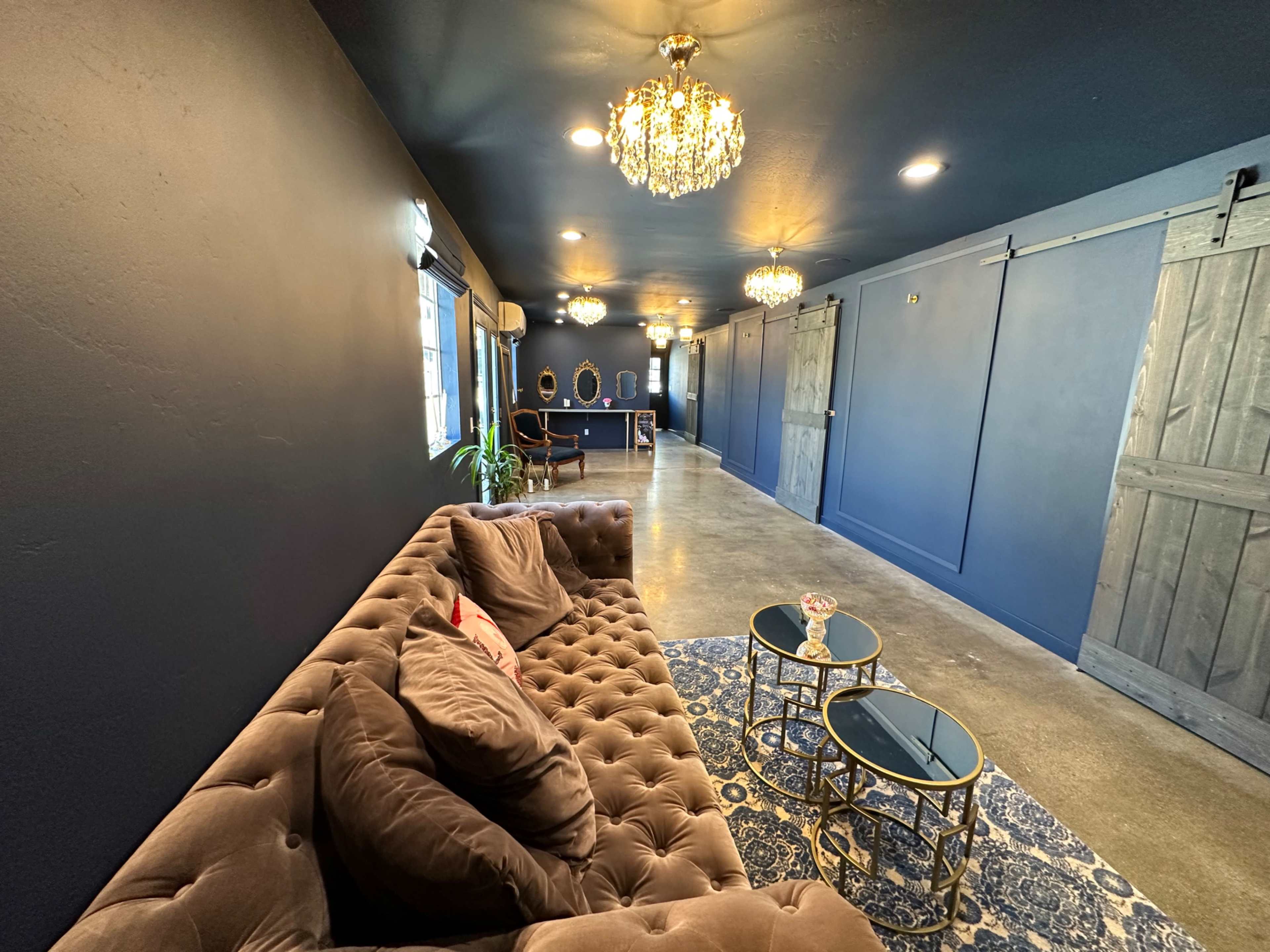 A modern hallway with a brown tufted sofa, decorative mirrors, two small gold tables, and pendant lighting against dark blue walls.