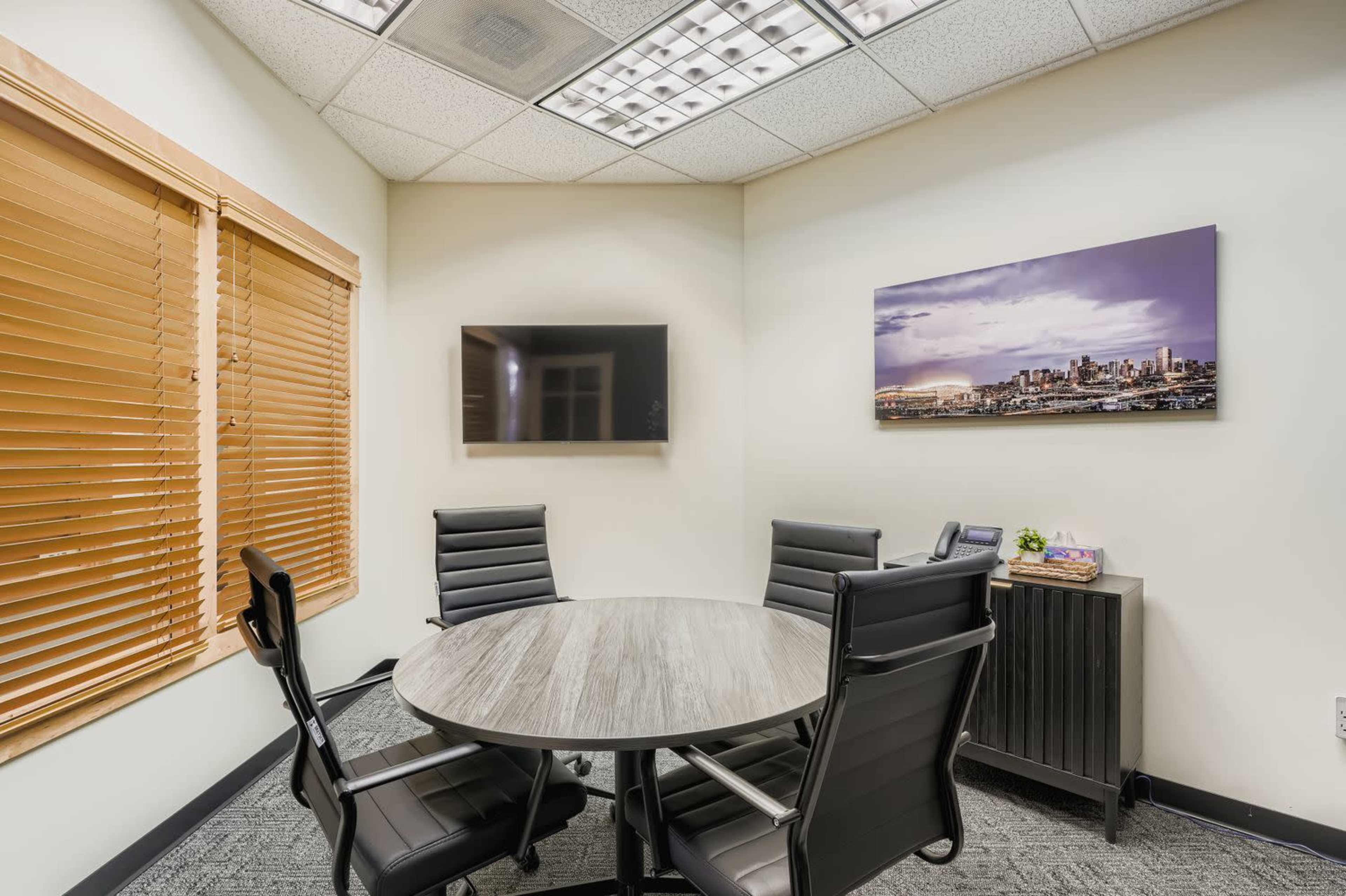 A small conference room features a round table surrounded by four black chairs, a wall-mounted television, and a window with wooden blinds.