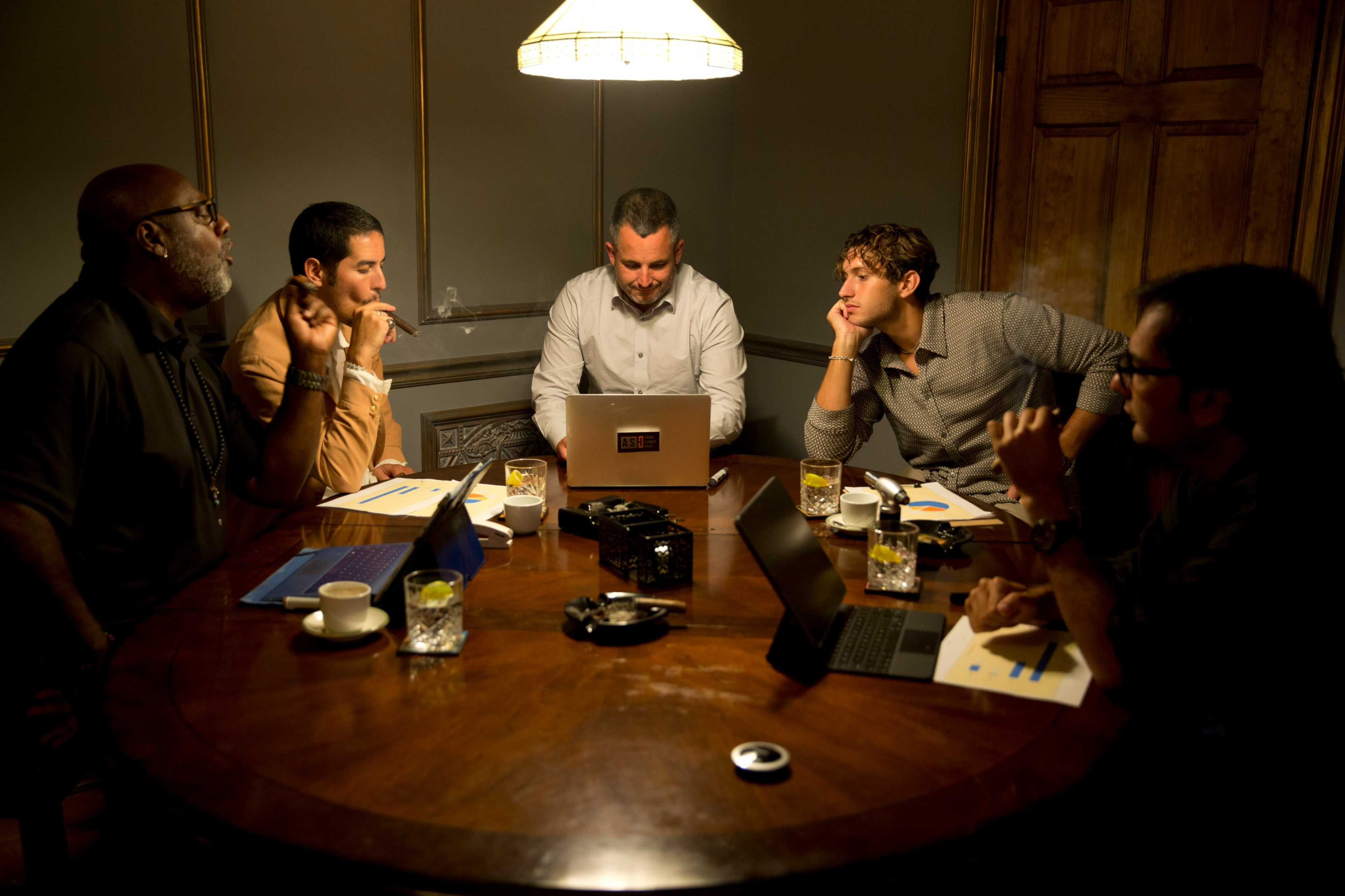 A group of five men is seated around a large wooden table, engaged in conversation while looking at a laptop in a dimly lit room.
