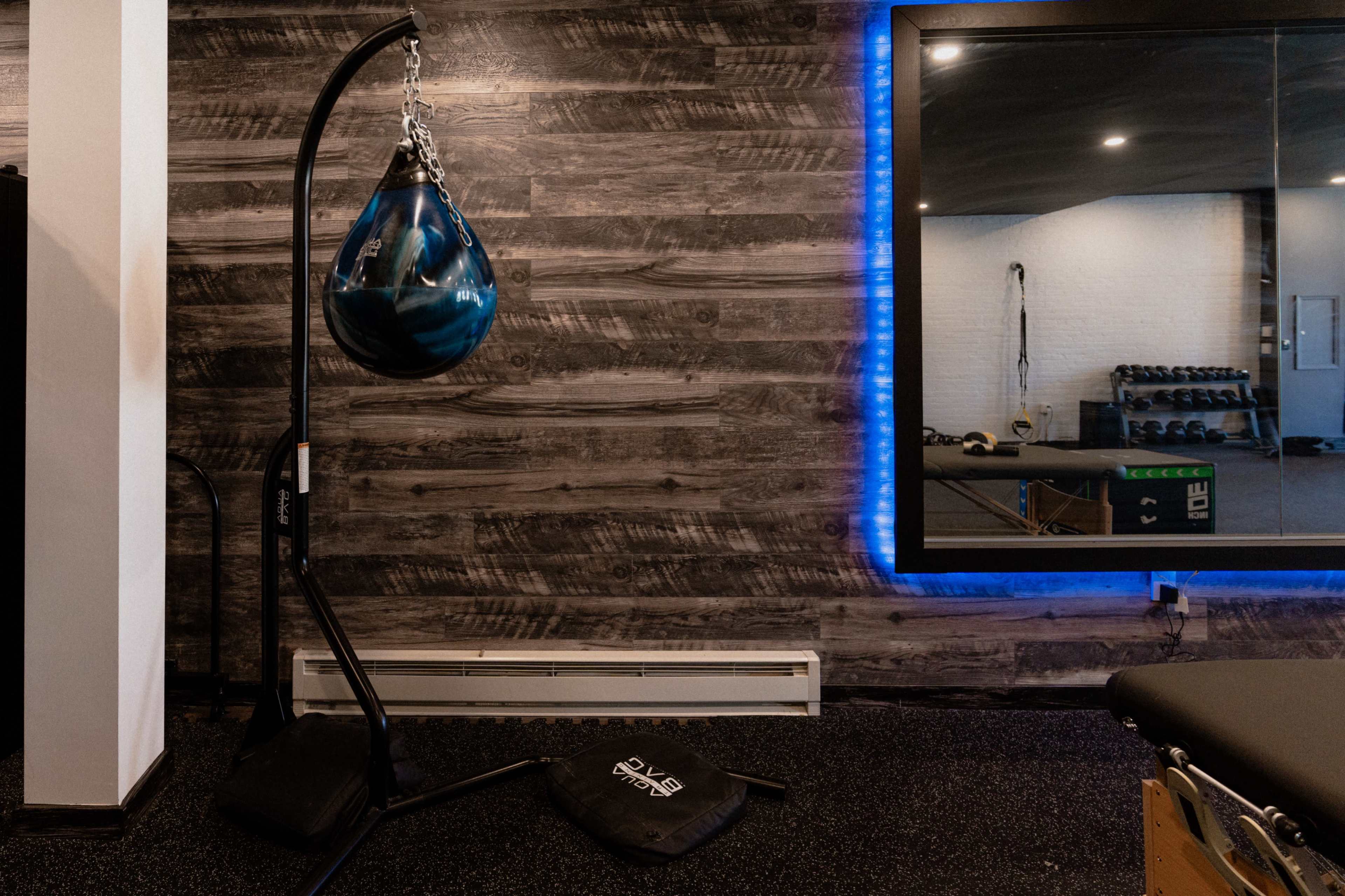 A gym interior featuring a heavy punching bag hanging from a stand, a large mirror reflecting gym equipment, and a textured wooden wall.