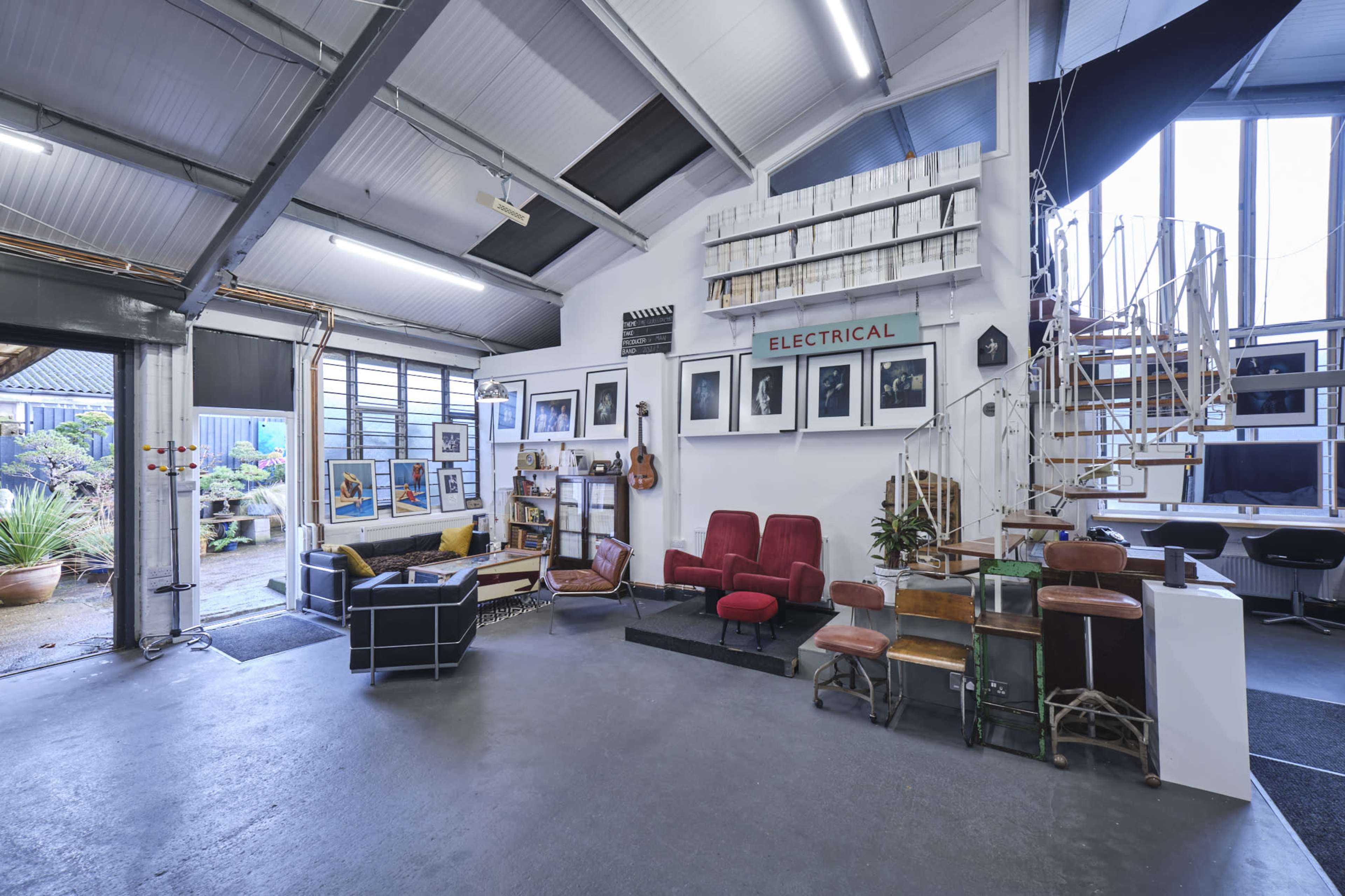 A spacious, industrial-style room featuring various seating arrangements, artwork on the walls, and a spiral staircase, with large windows allowing natural light to enter.