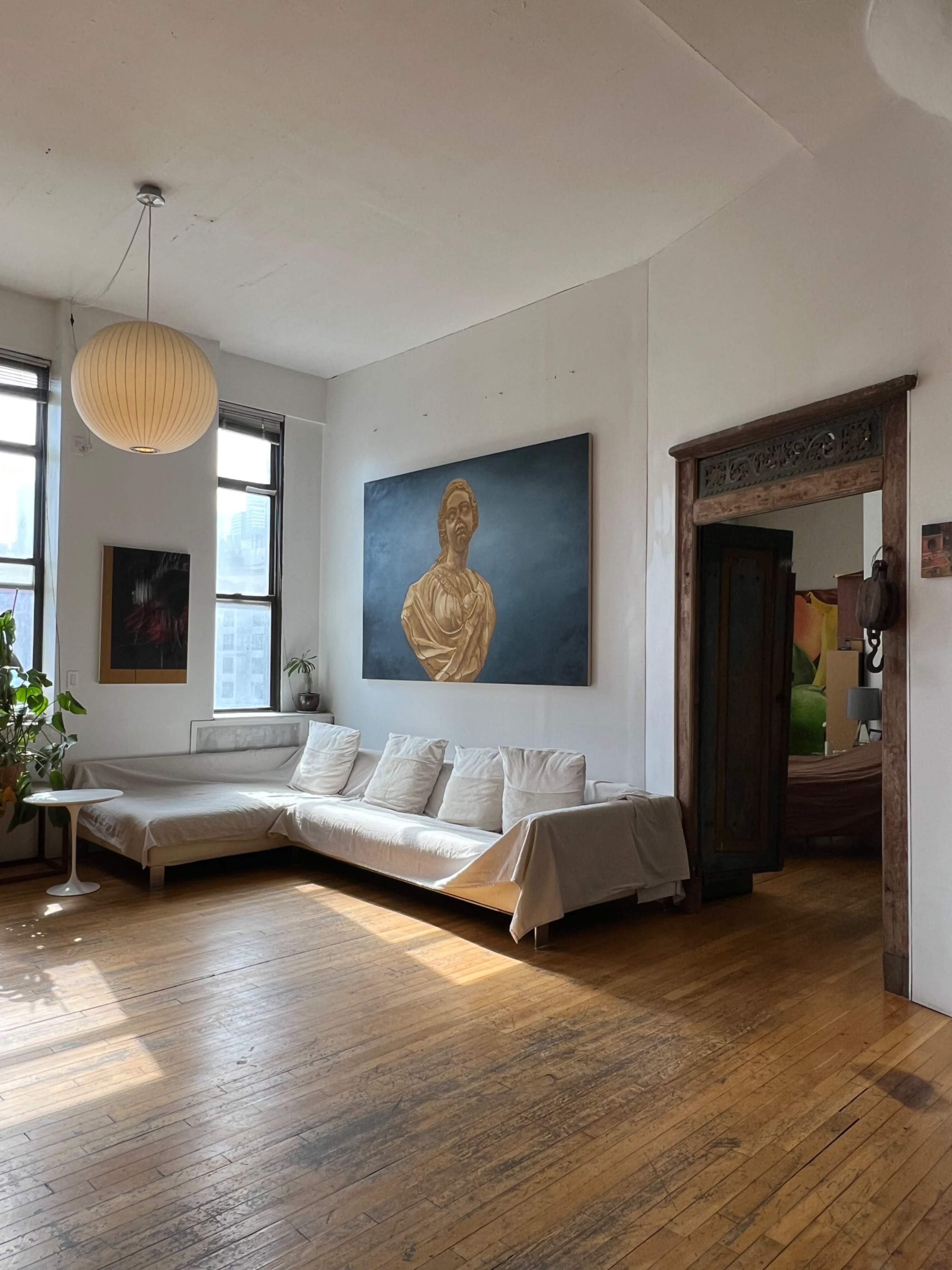 The room features a large white sofa, a prominent gold painting on the wall, and wooden flooring with large windows allowing natural light.