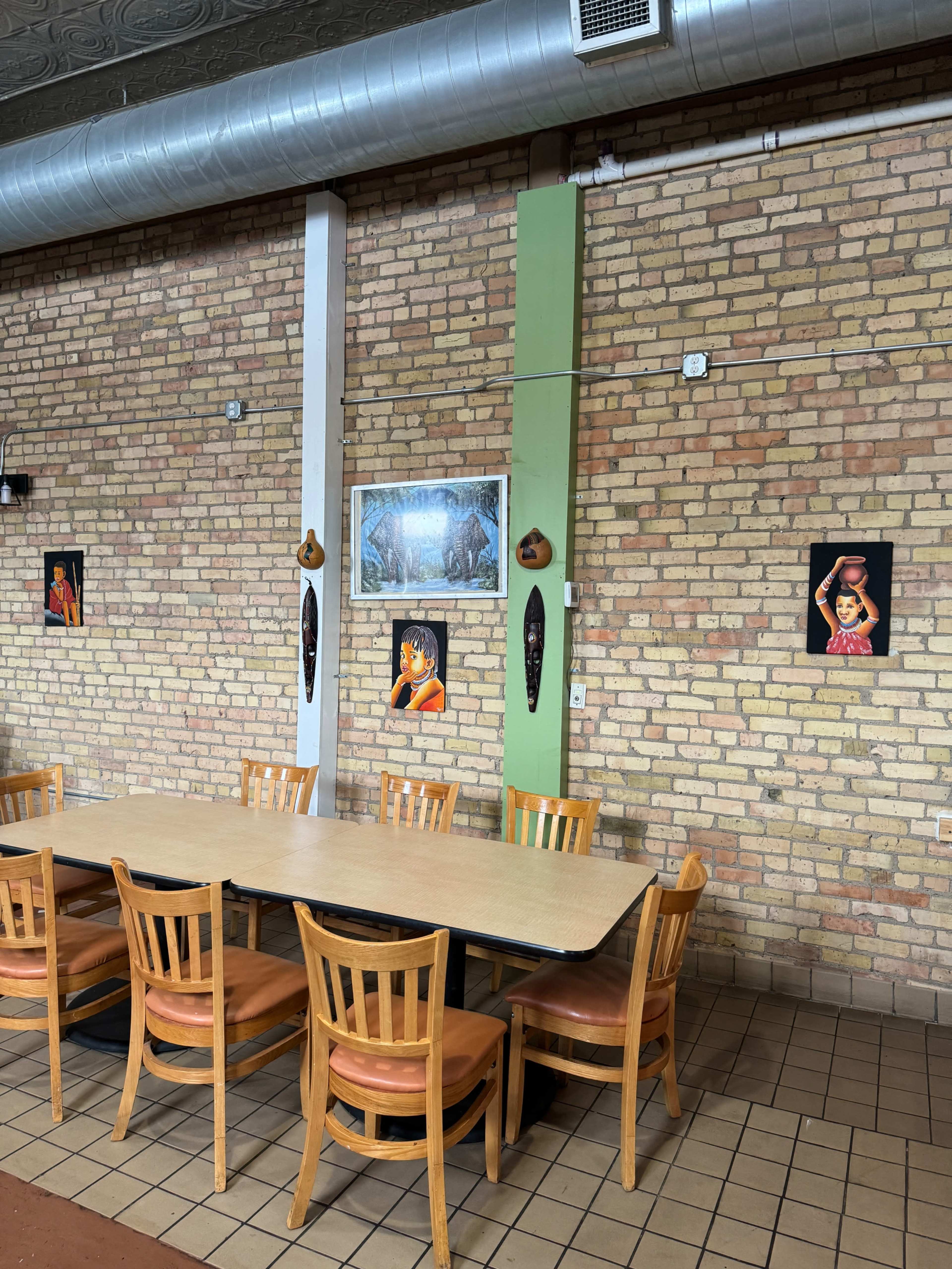 The image shows a long wooden table with chairs in a dining area, surrounded by brick walls adorned with framed pictures and decorative masks.