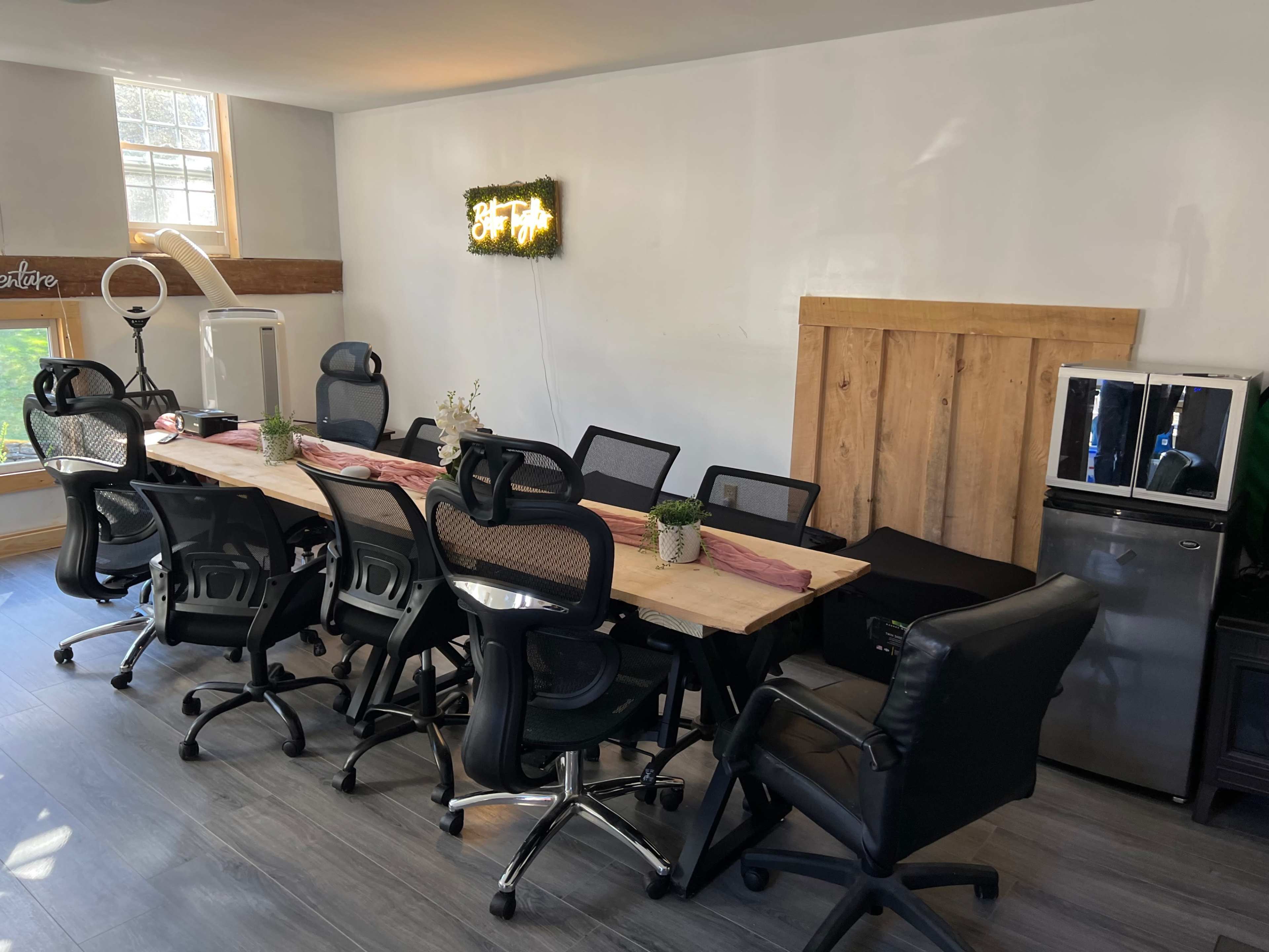 A modern conference room with a long wooden table surrounded by black office chairs, a mini fridge, and a neon sign on the wall.