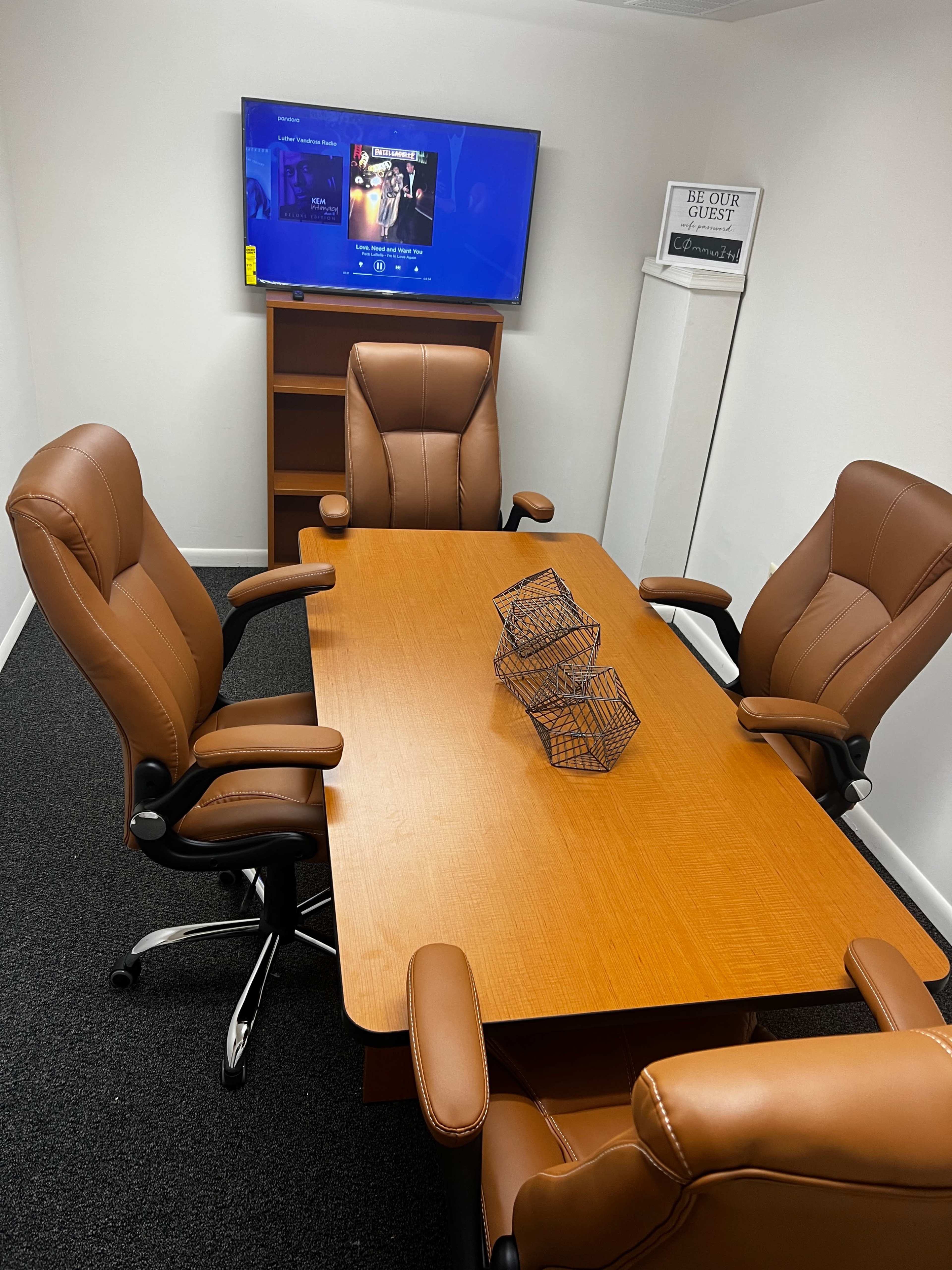 A small conference room features a wooden table surrounded by four brown leather office chairs, with a television mounted on the wall displaying a menu and two wire baskets on the table.