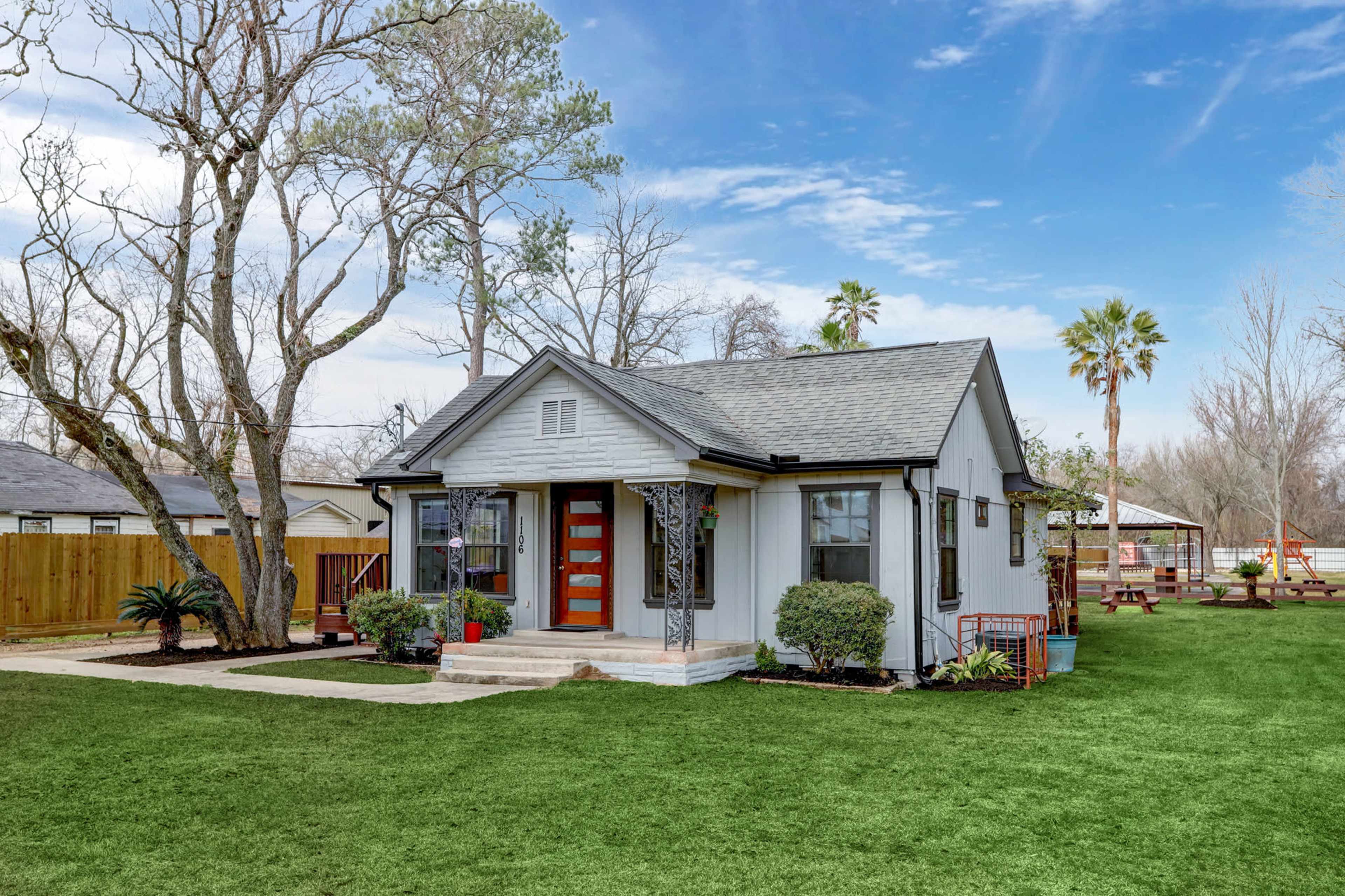 A small, modern single-story house with a gray exterior and bright orange doors sits in a lush green yard surrounded by trees and a play area.