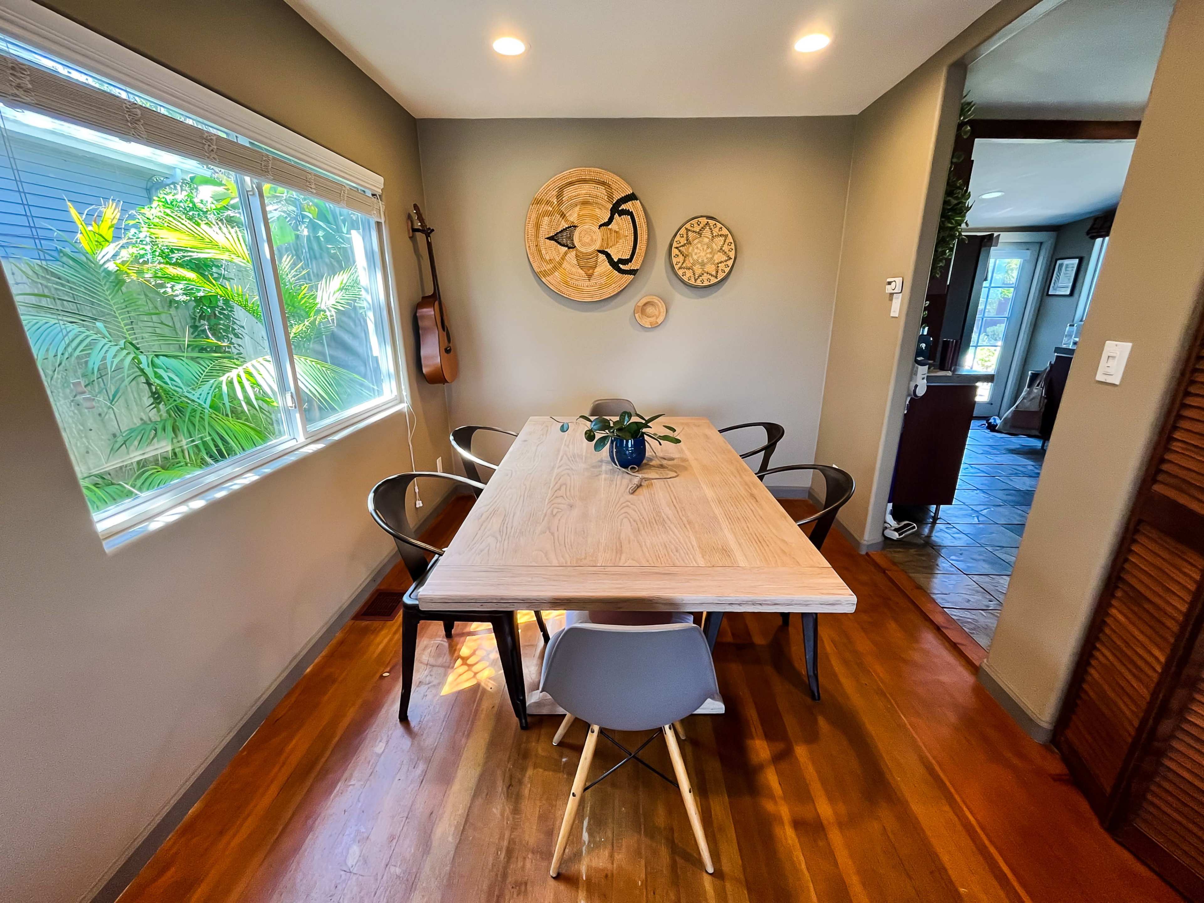 A modern dining area features a wooden table surrounded by black and white chairs, with decorative wall art and a window overlooking plants.