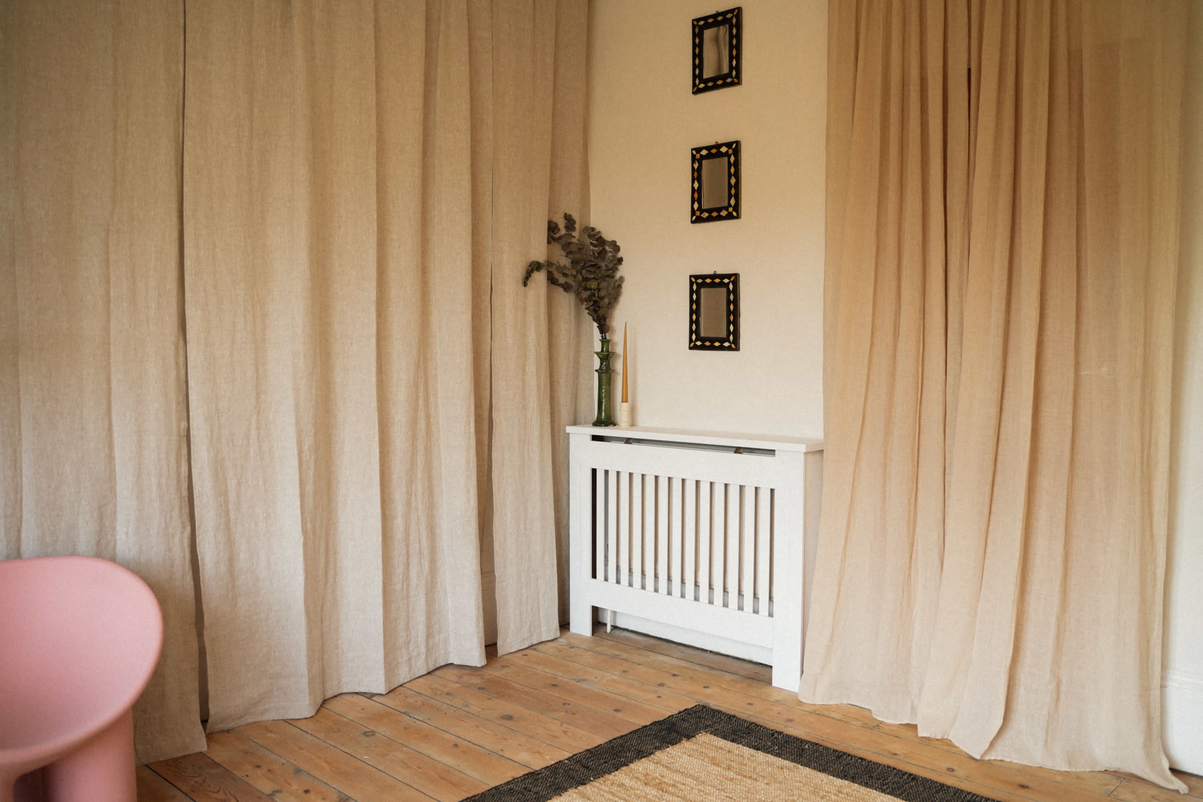 The image shows a corner of a room with beige curtains, a white radiator cover, and three framed mirrors hanging on the wall above a small vase with dried flowers.