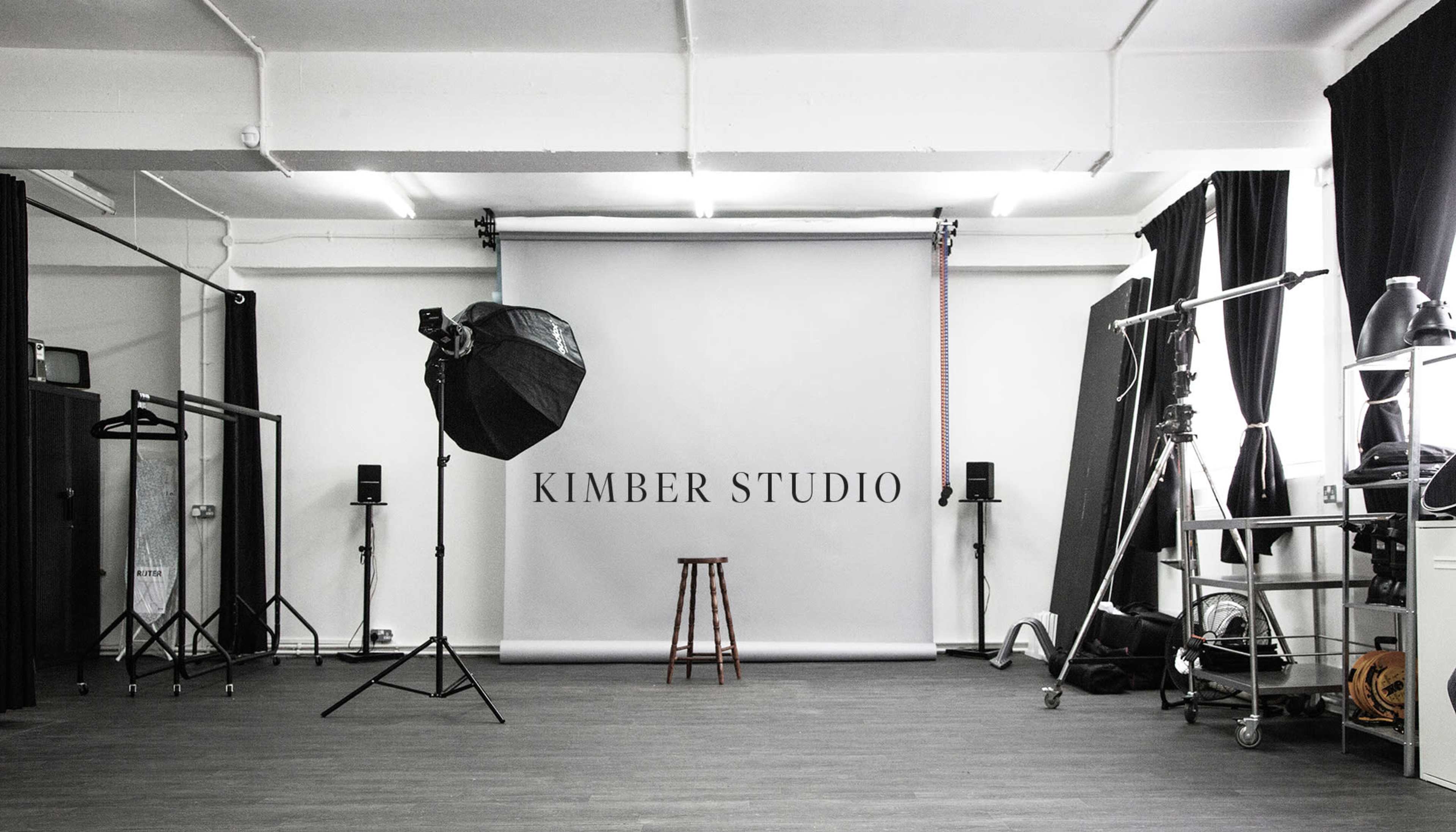 A photography studio with a plain backdrop, a stool, lighting equipment, and various studio furniture arranged neatly.