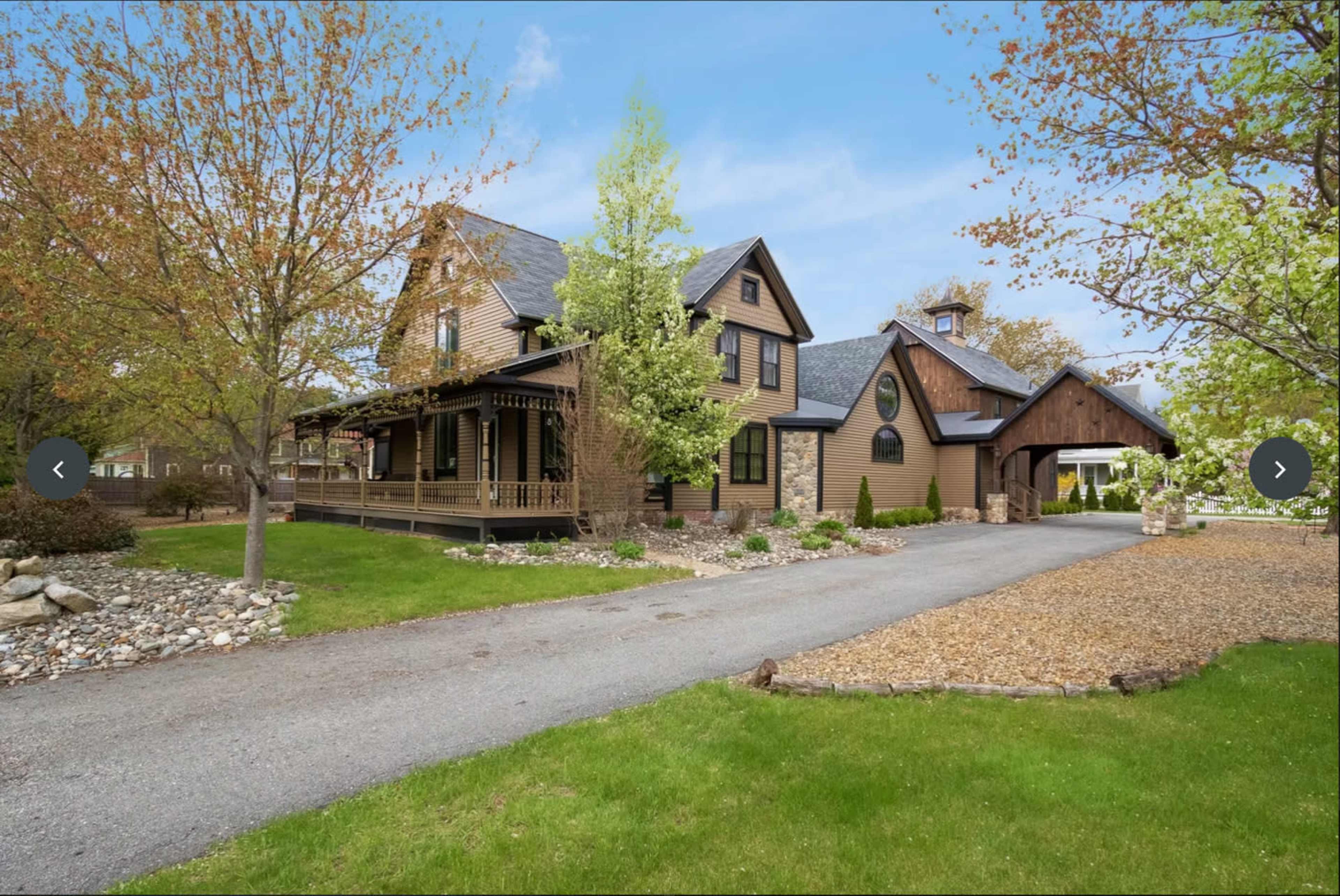 A two-story house with a mix of wood and stone exterior, situated beside a gravel driveway and landscaped yard with trees and shrubs.
