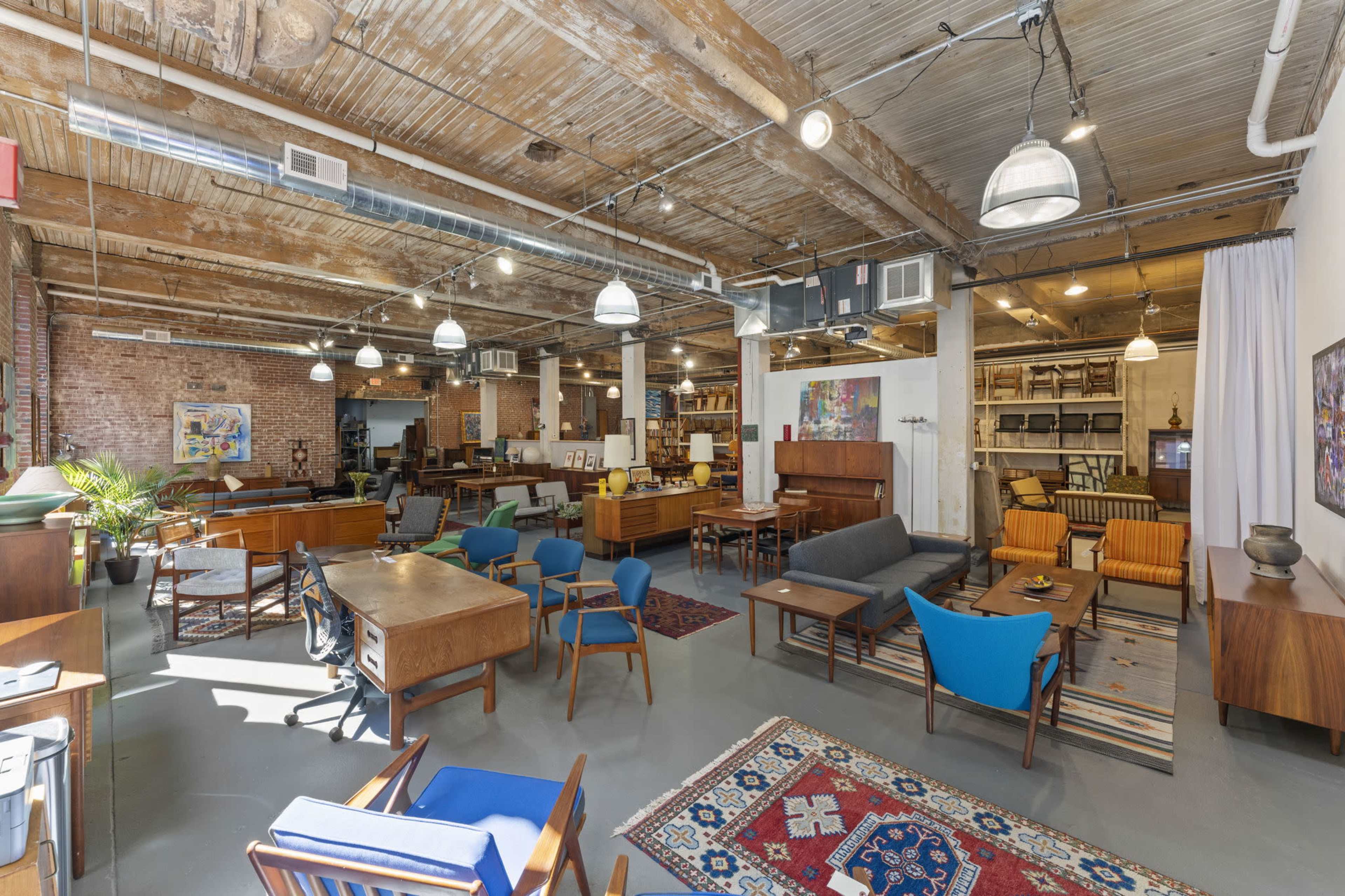 A spacious furniture showroom featuring a variety of Mid-Century modern pieces, with exposed wooden beams and industrial lighting.