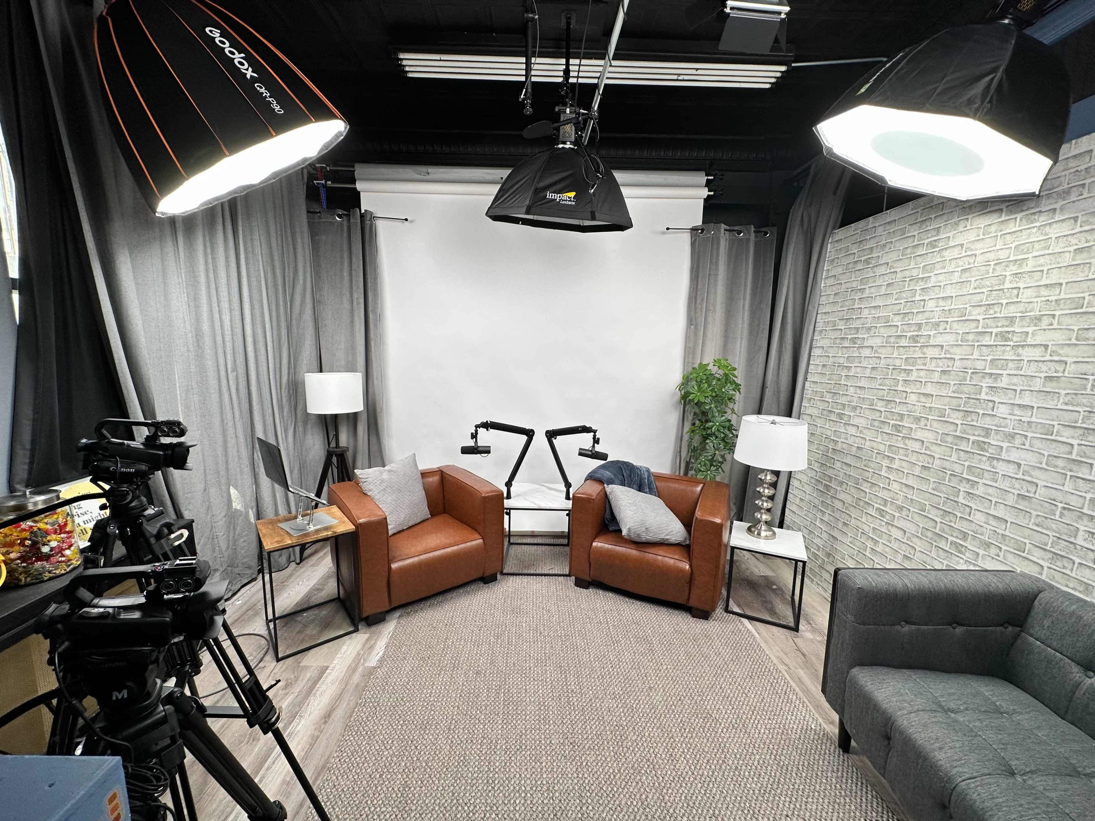 A studio setup with two brown leather chairs facing a white backdrop, flanked by lamps and a potted plant, with a camera and colorful snacks on a table in the foreground.
