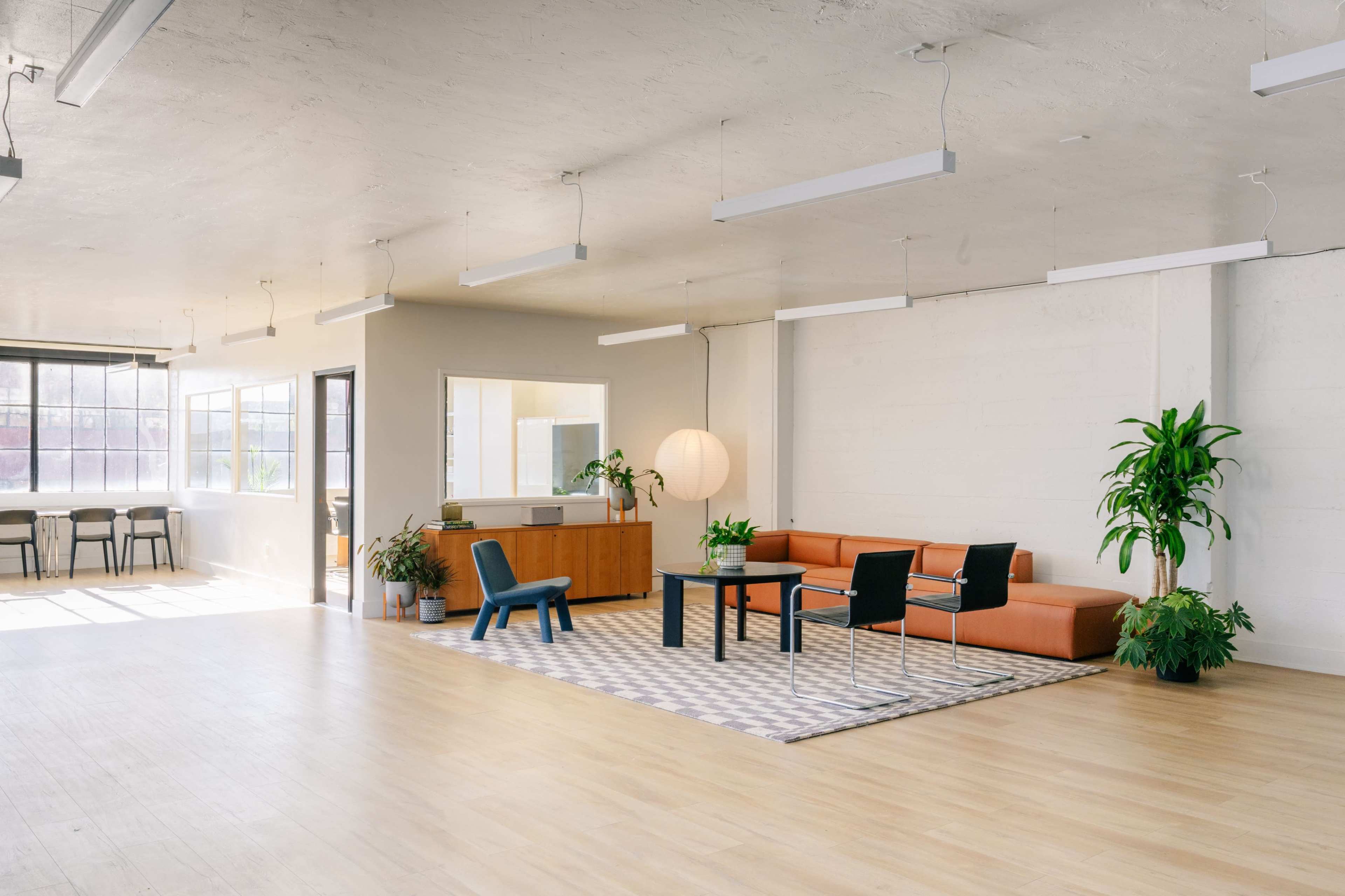 The image shows a spacious, modern office interior featuring a terracotta sofa, a coffee table, and various plants, with large windows allowing natural light.