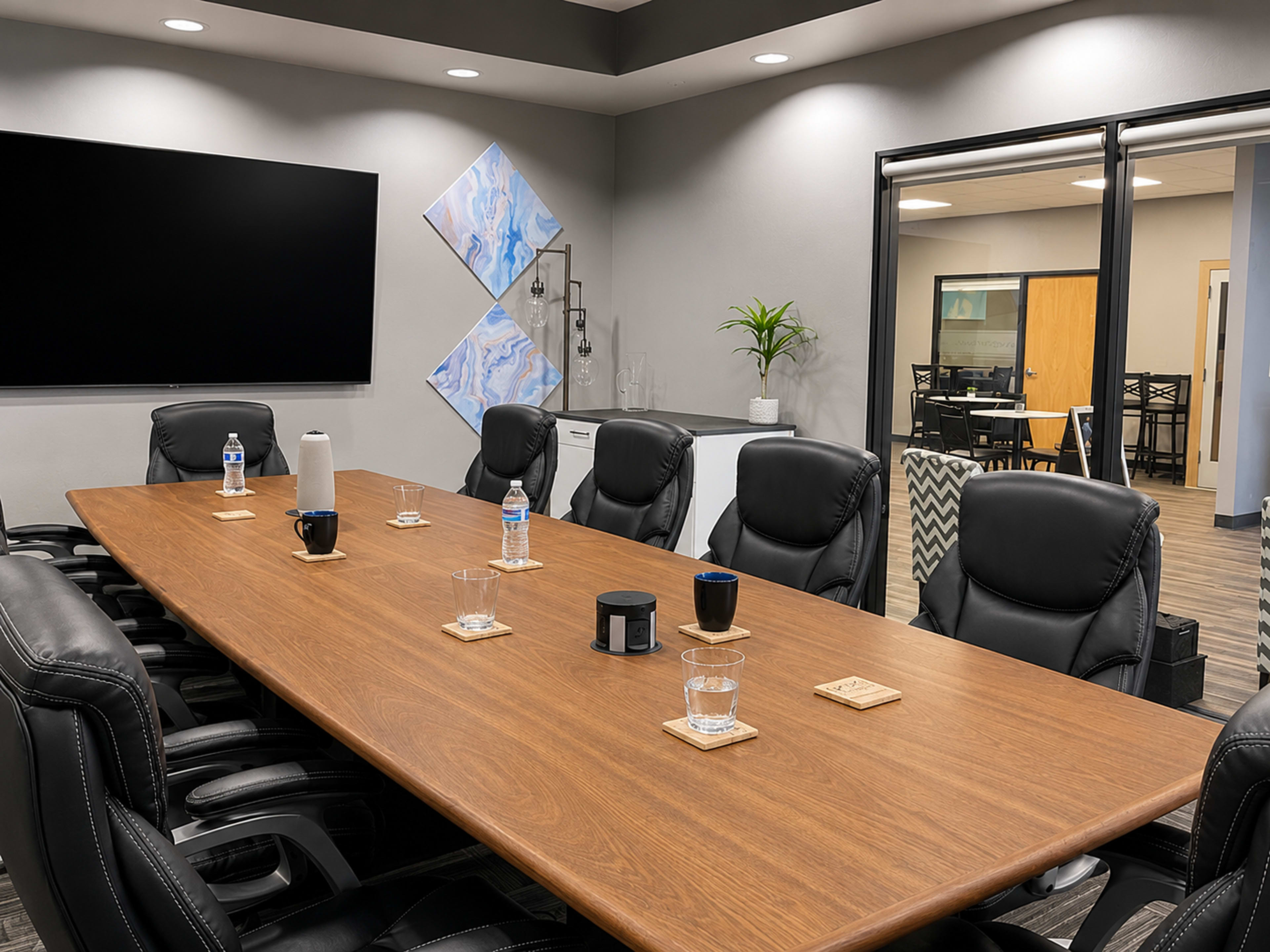 A large conference room features a long wooden table surrounded by black leather chairs, with a television screen on one wall and abstract art pieces on another.