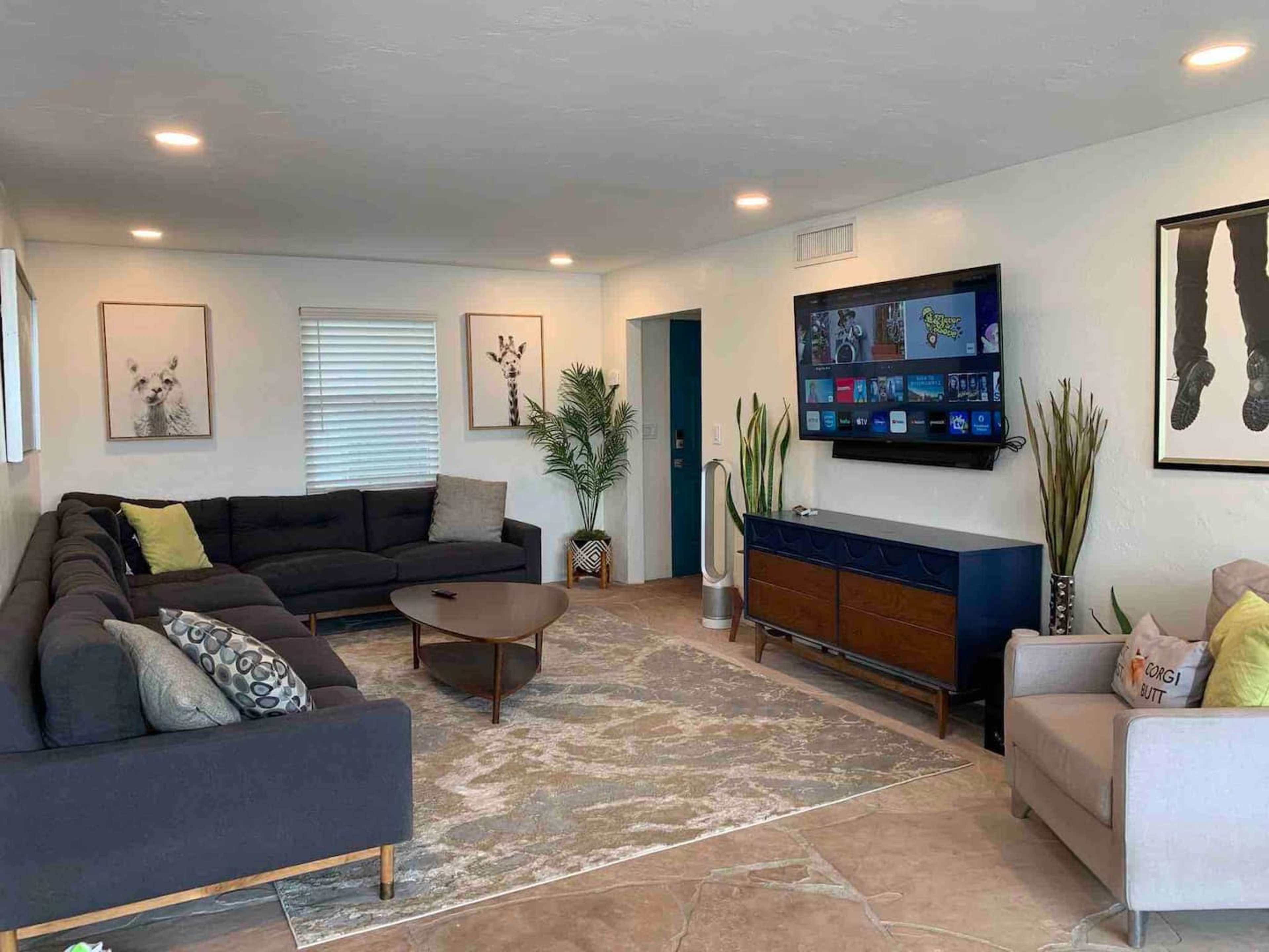 A modern living room features a large sectional sofa, a wall-mounted television, and decorative plants against a light-colored wall.