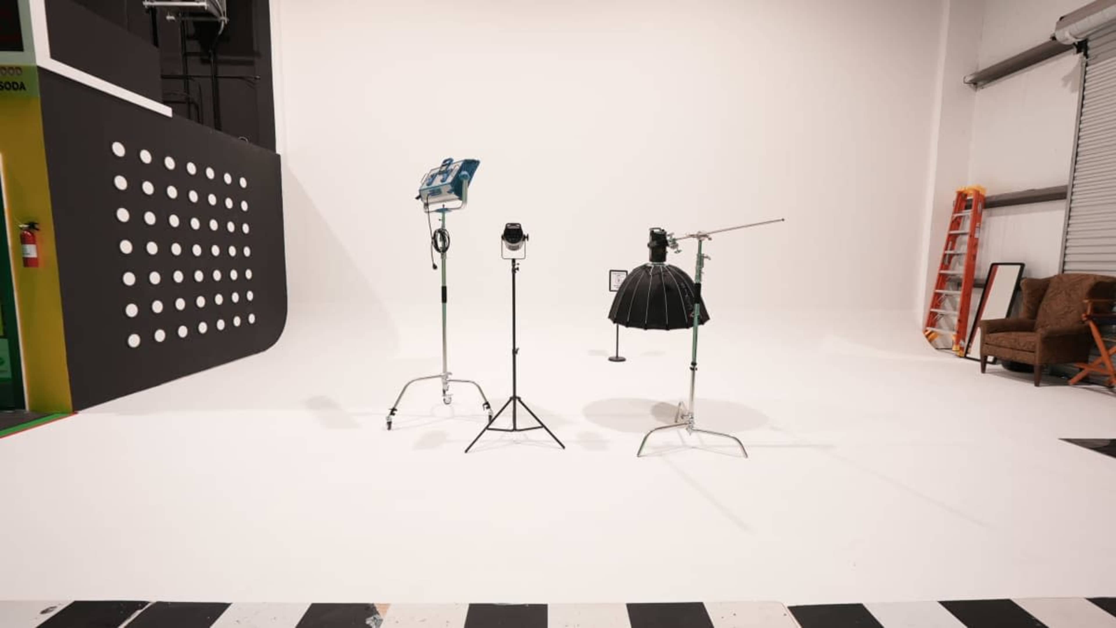 The image shows a photography studio with lighting equipment, a ladder, and a brown couch against a white backdrop.