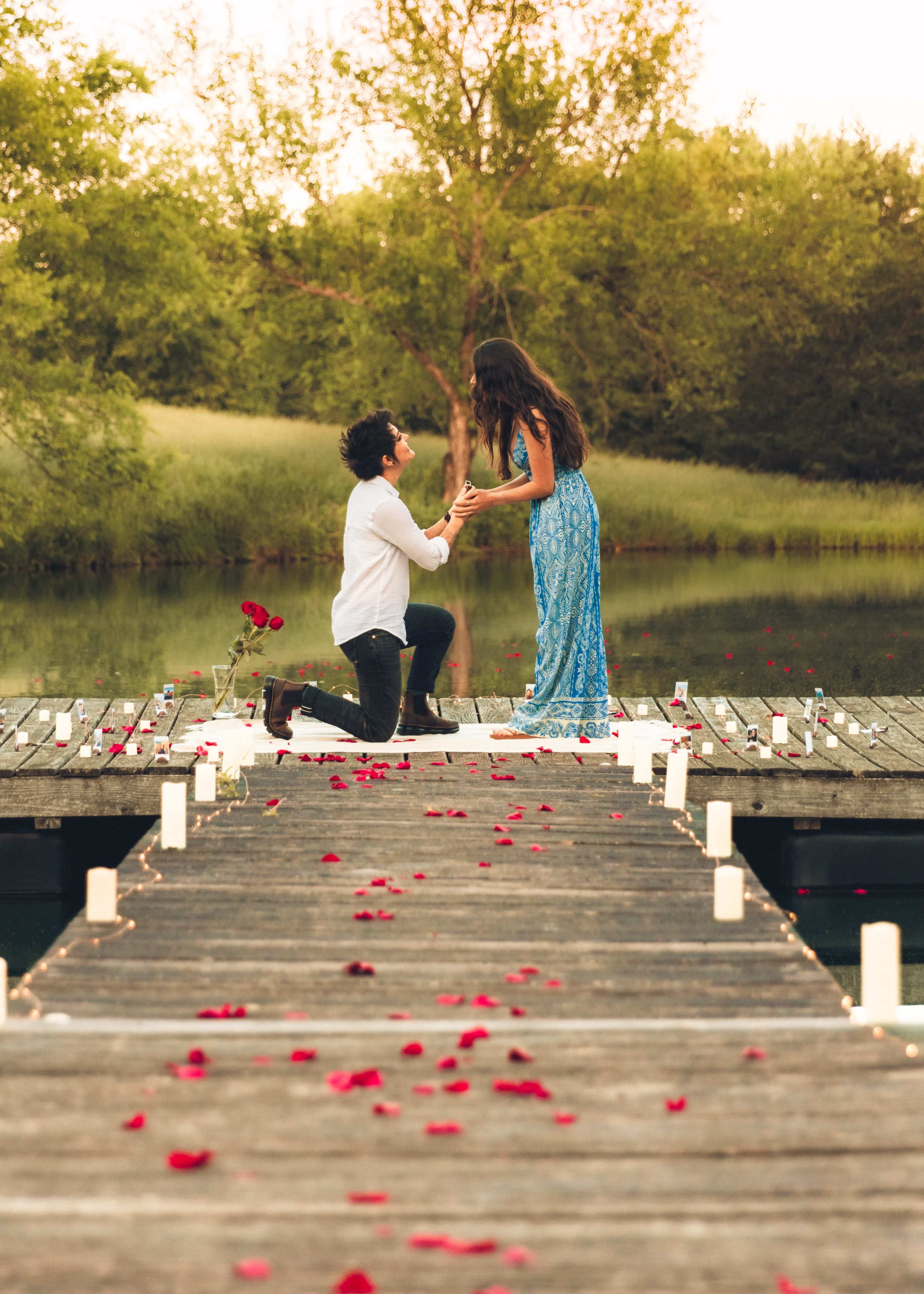 A man kneels on a dock sprinkled with rose petals while proposing to a woman in a blue dress, with candles lining the path beside a serene body of water.