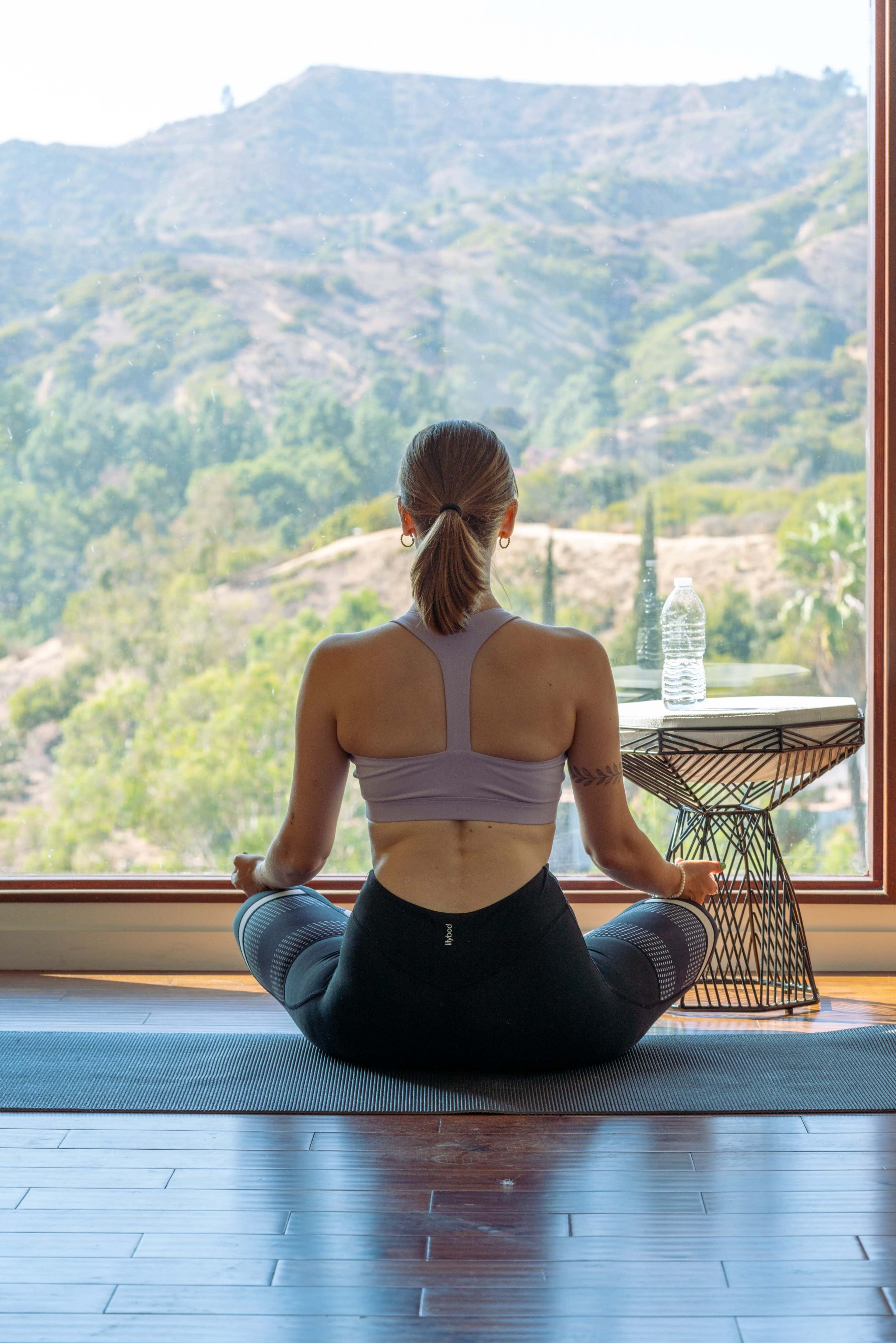 A person sits cross-legged on a mat in a room with a large window that overlooks a mountainous landscape.