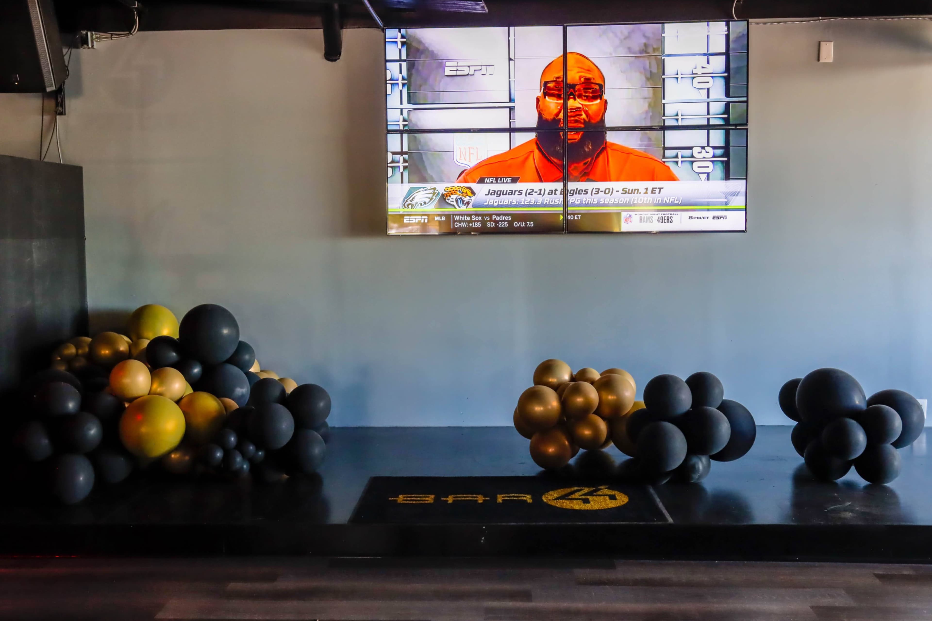 A television displaying a sports broadcast is mounted on a black wall, with clusters of black, gold, and white balloons arranged on the floor below.