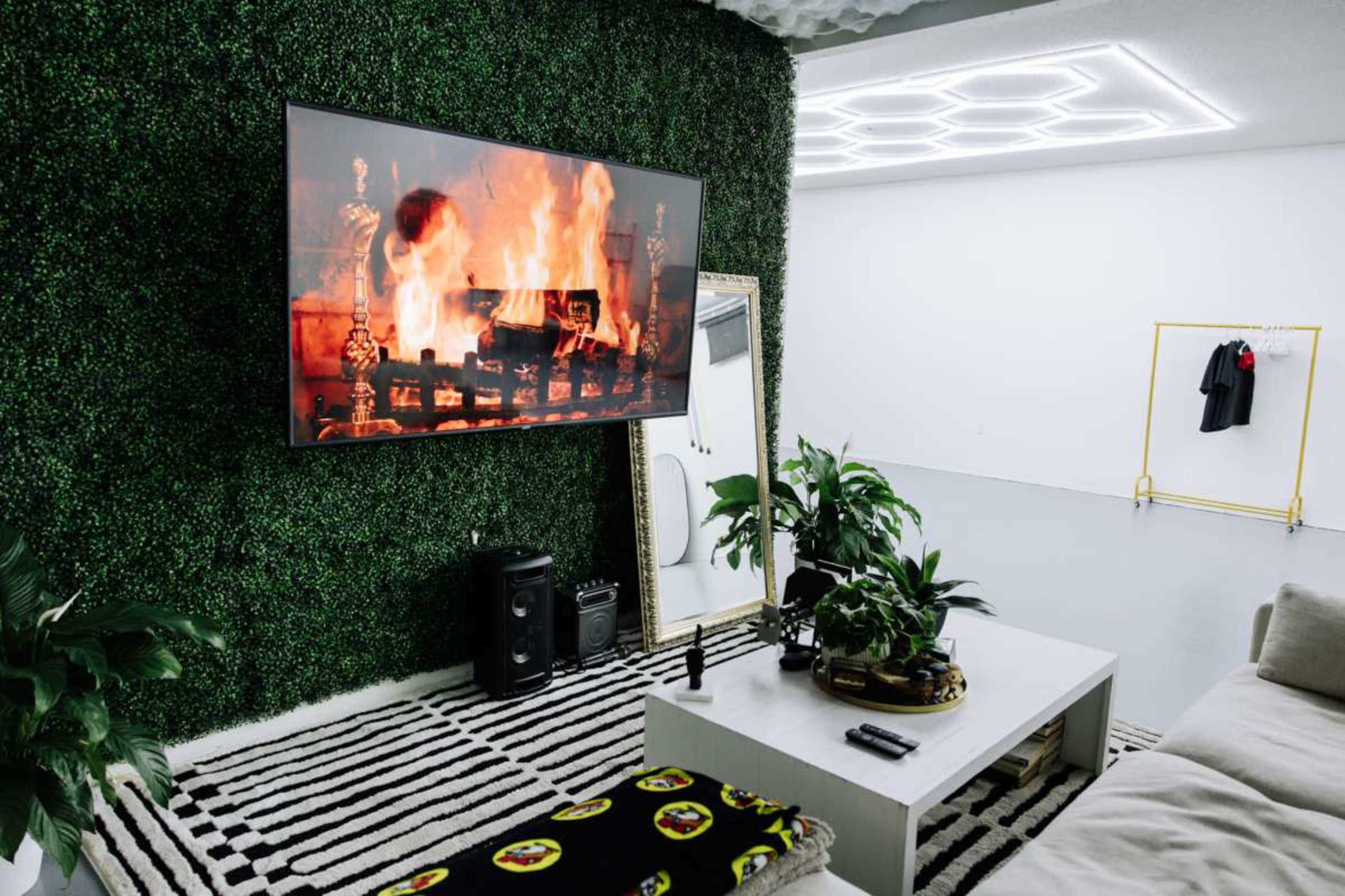 The image shows a modern living room with a large TV displaying a fireplace, surrounded by greenery on the walls and a striped rug on the floor.