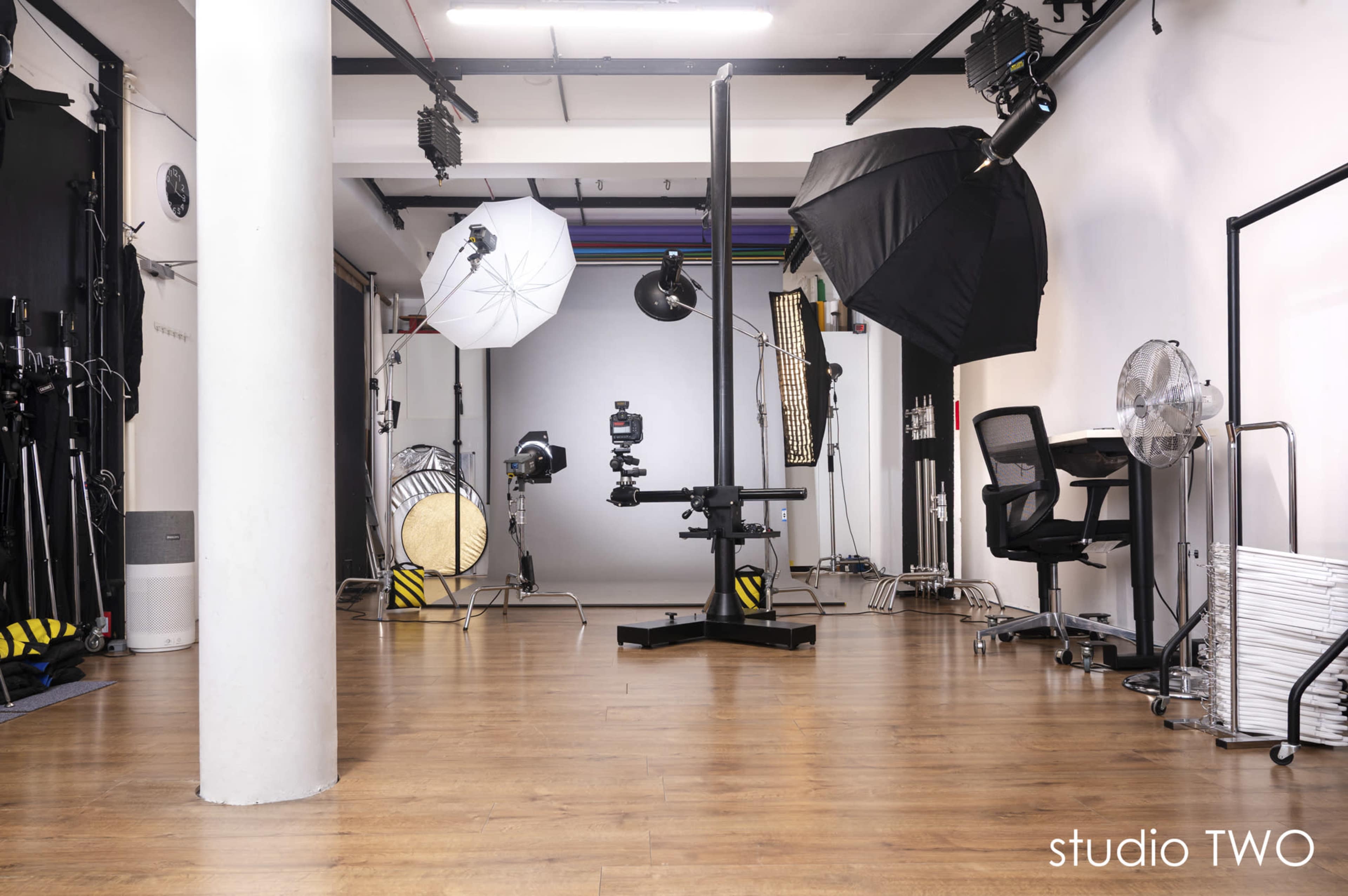 The image shows a photography studio equipped with various lighting setups, a camera on a stand, and a work desk placed in the corner.