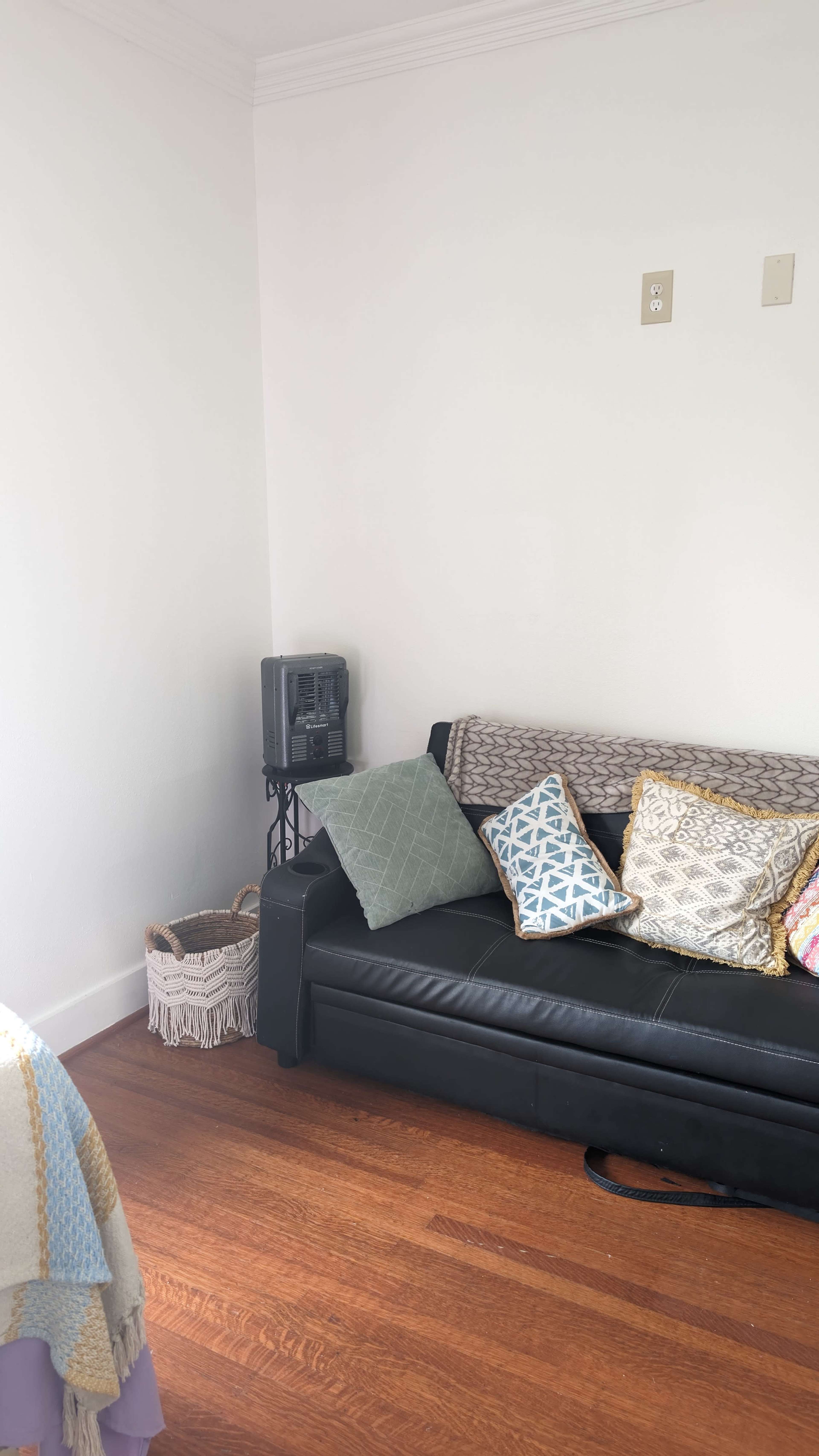 A black sofa with various patterned cushions is positioned against a white wall in a room with wooden flooring.