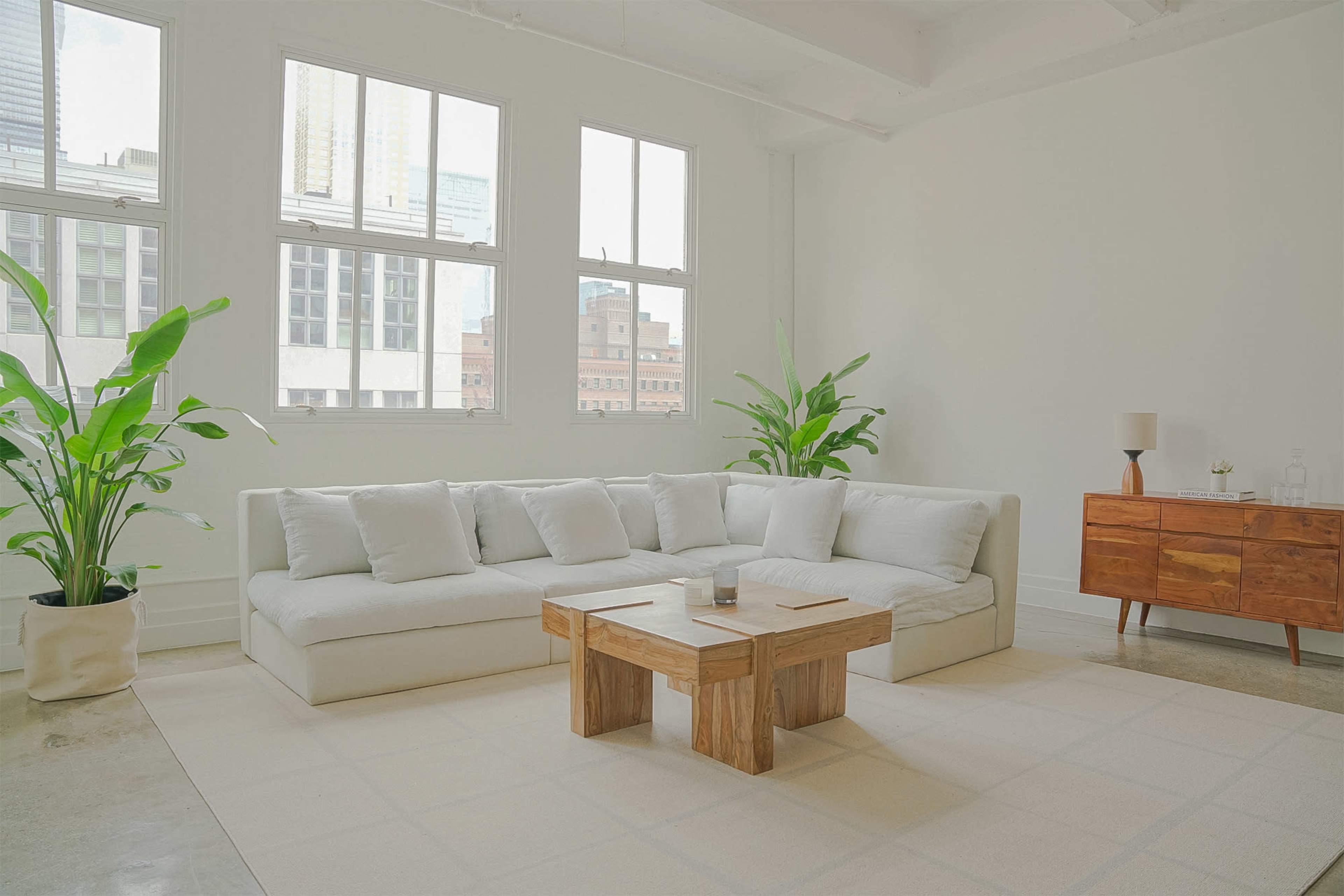 A light and airy living room features a large, L-shaped white sofa, a wooden coffee table, and several indoor plants beside a mid-century modern chest of drawers.