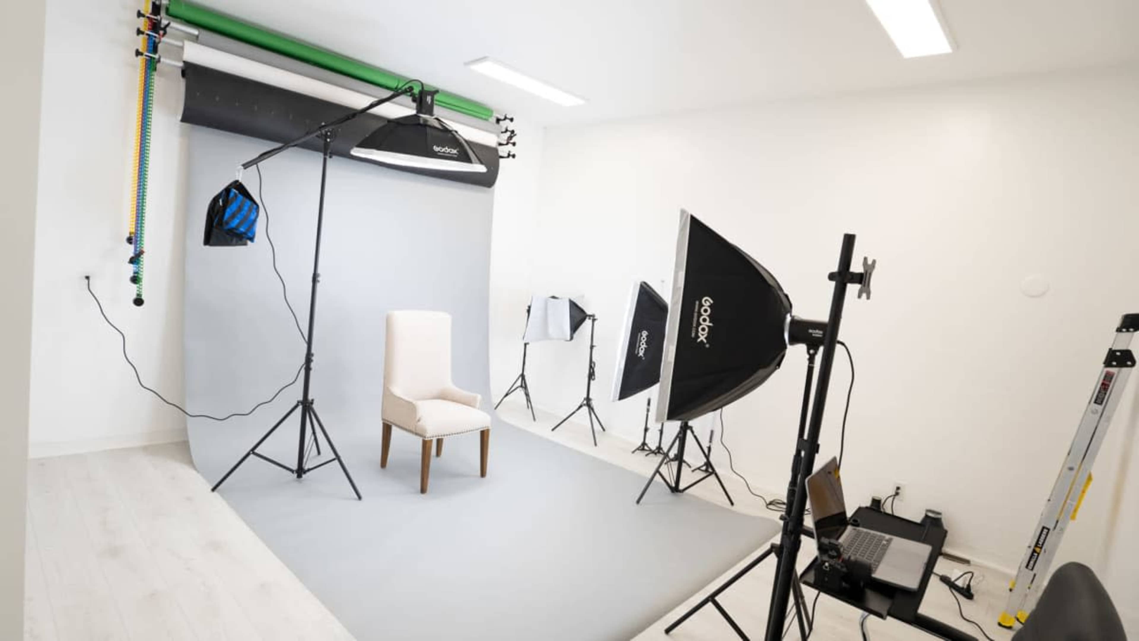The image shows a photography studio set up with a simple chair on a gray backdrop, surrounded by various lighting equipment and a ladder.