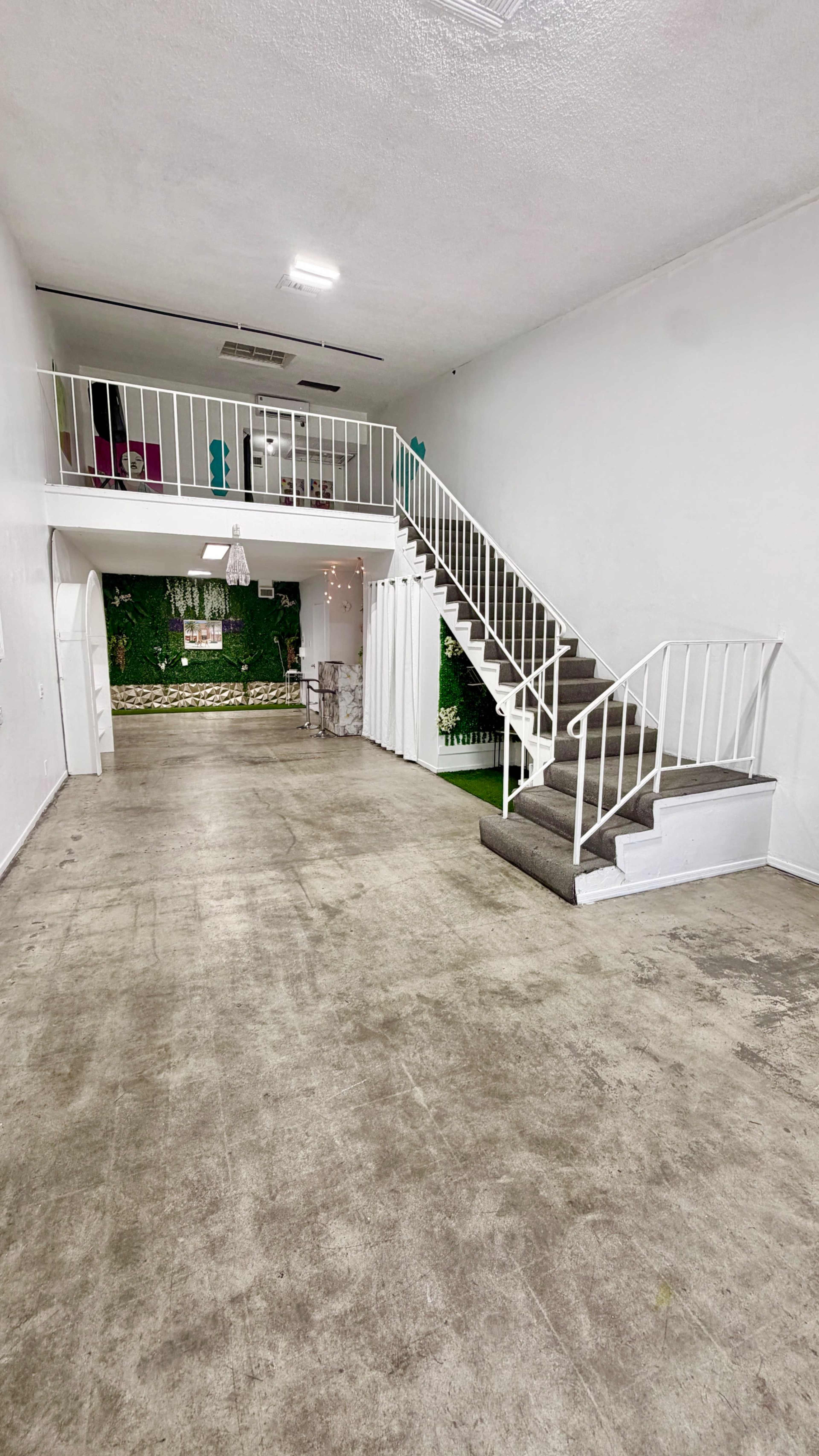 The image shows an empty, open space with a staircase on the right leading to a second level, and a wall with greenery in the background.