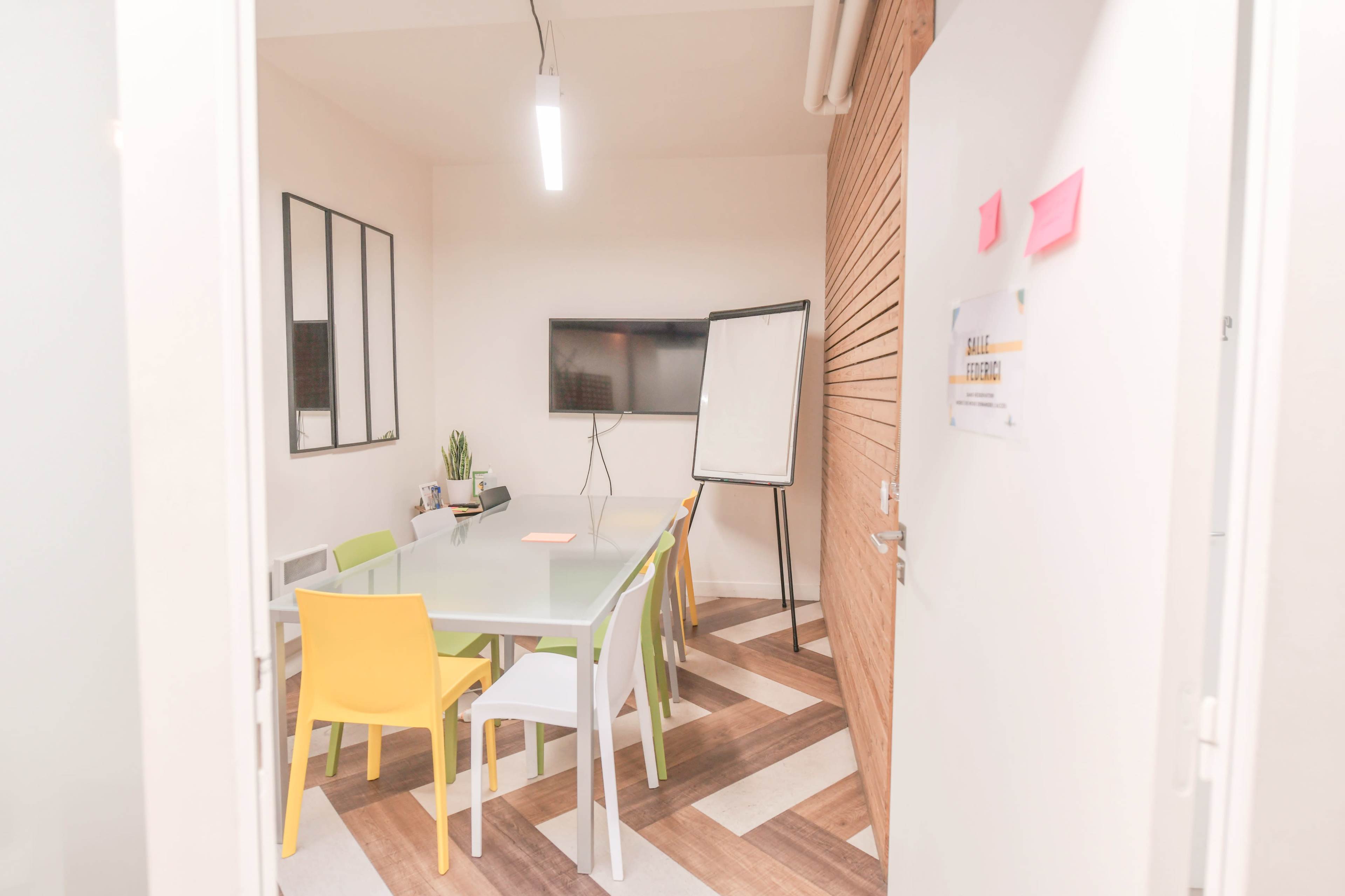 A brightly lit meeting room features a glass table surrounded by colorful chairs, a wall-mounted TV, and a whiteboard.