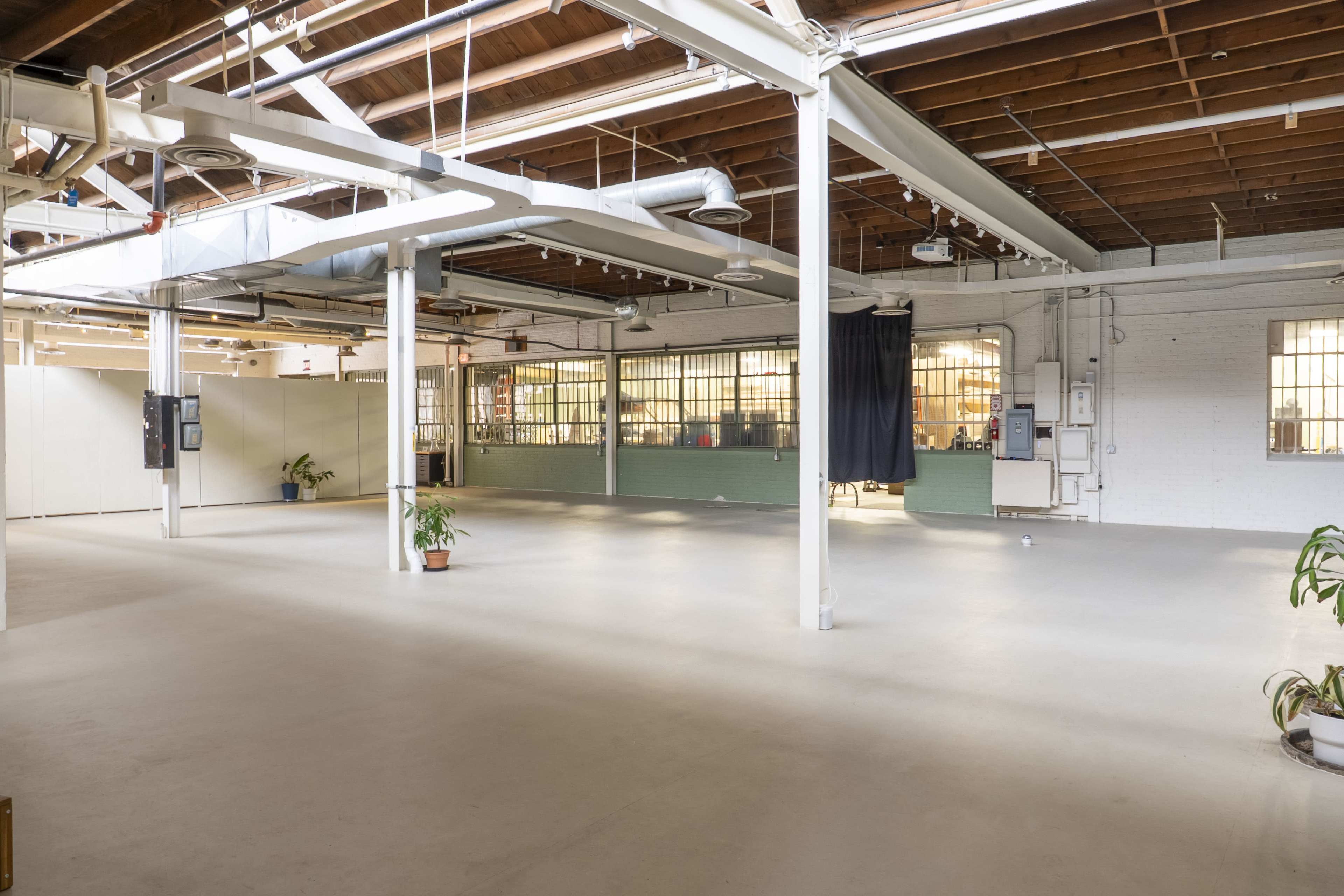 The image shows a spacious, empty industrial-style room with high ceilings, exposed beams, and large windows.