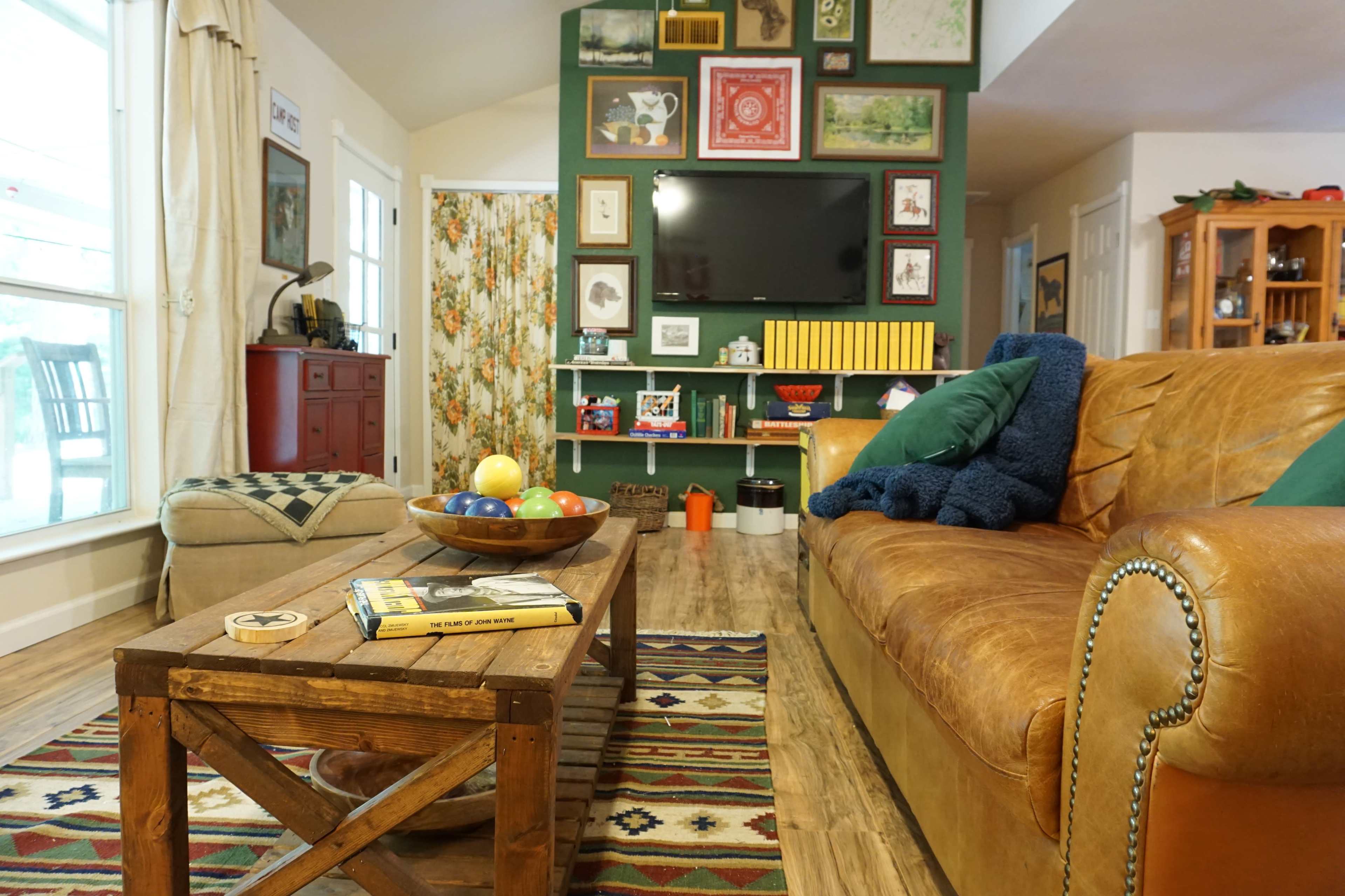 The living room features a brown leather sofa, a wooden coffee table with a bowl of colorful balls, and a wall decorated with various framed artworks.