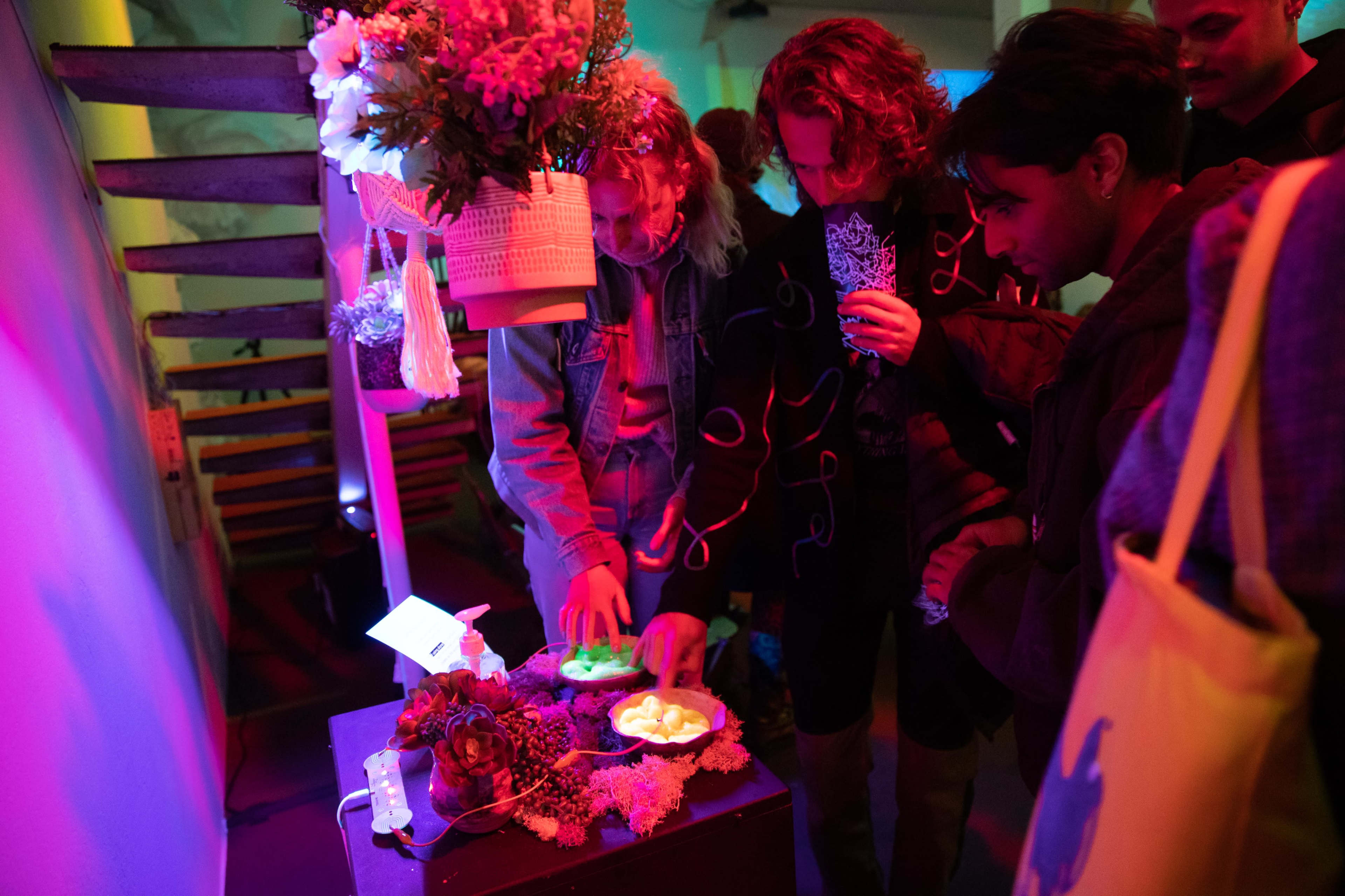 A group of three individuals examines a small display featuring flowers, lights, and a plate of objects in a dimly lit space.
