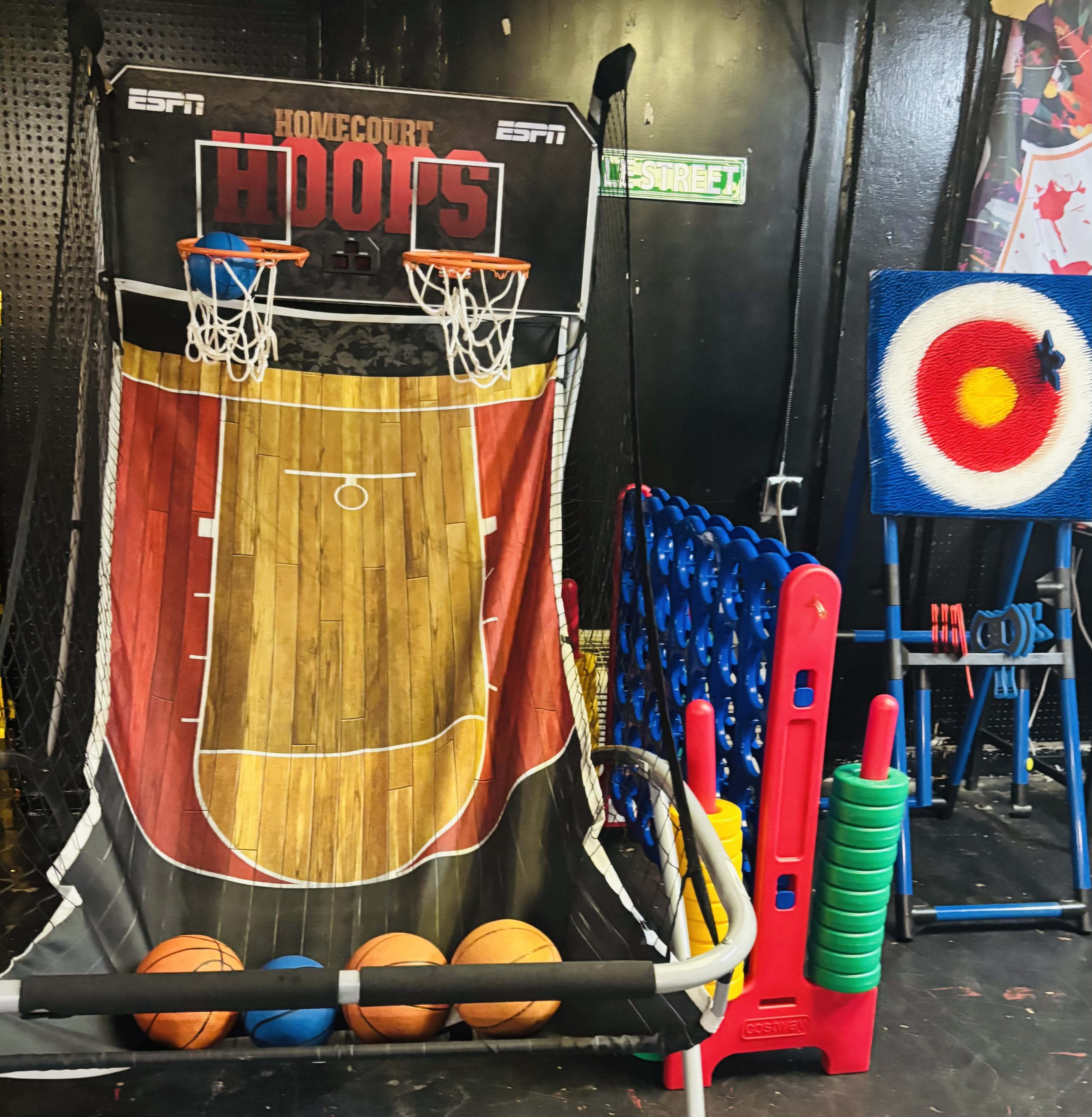 The image shows an arcade basketball game with a large backdrop featuring a basketball court, surrounded by colorful balls and a target shooting game on the right.