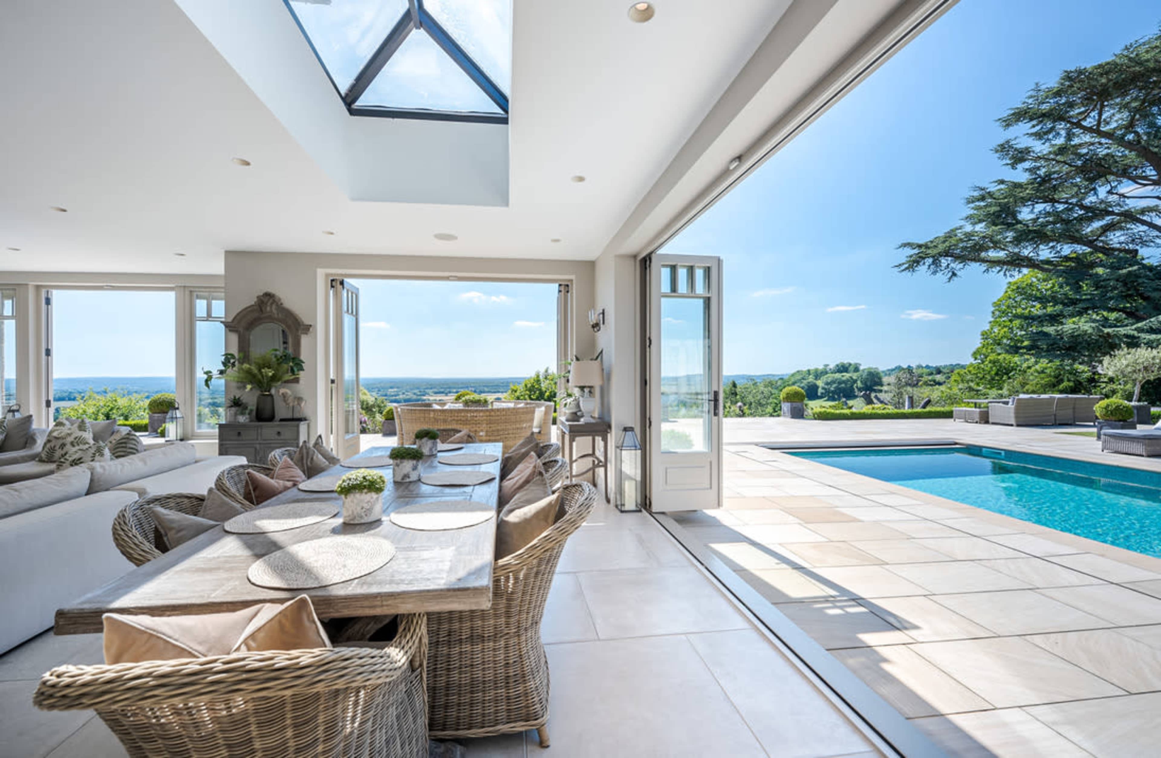 The image shows a modern dining area with a wooden table surrounded by wicker chairs, large glass doors leading to a patio with a swimming pool and a scenic view in the background.