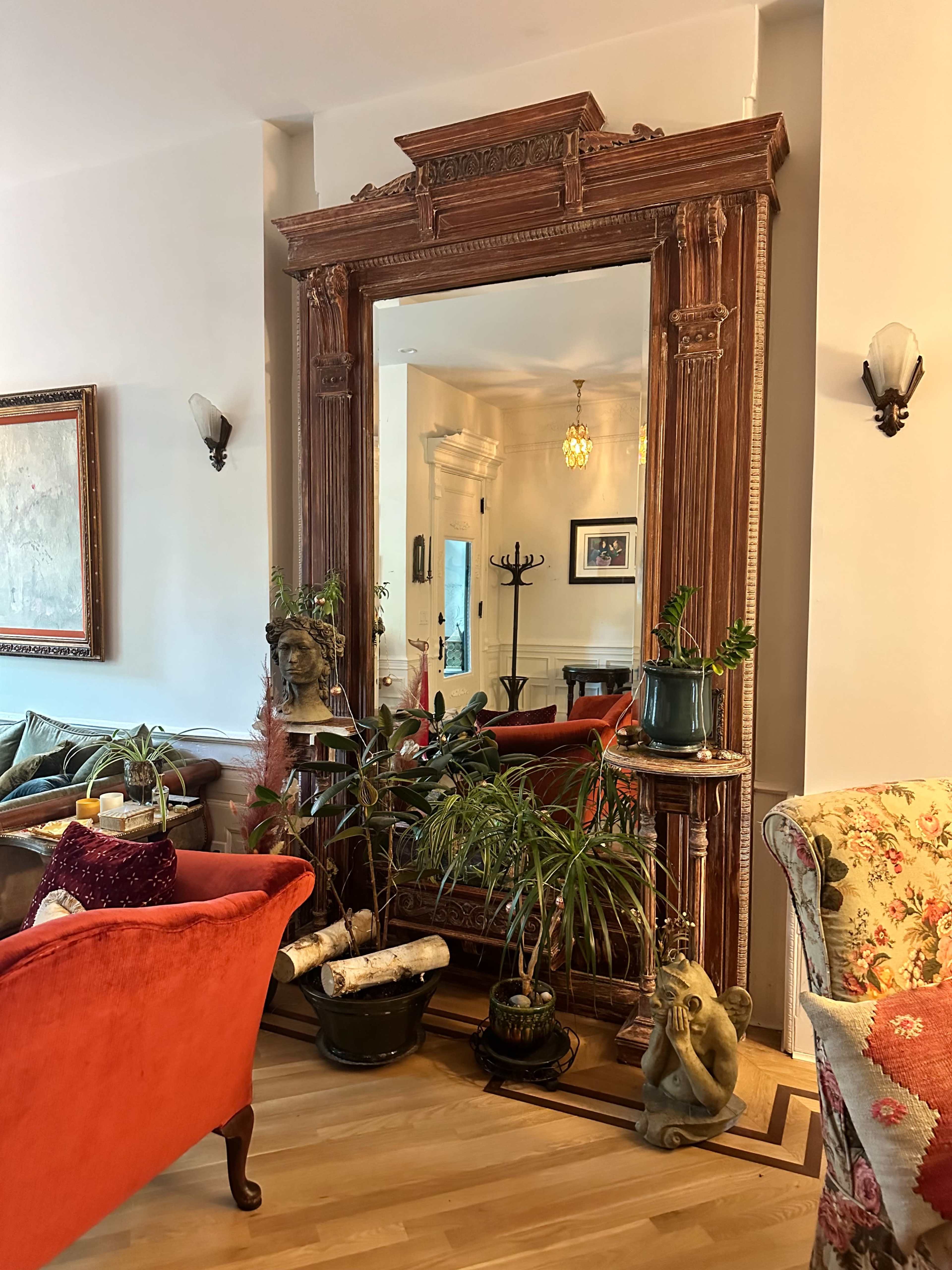 A large ornate mirror is framed by a decorative wooden border and surrounded by various plants and furniture in a cozy living room.