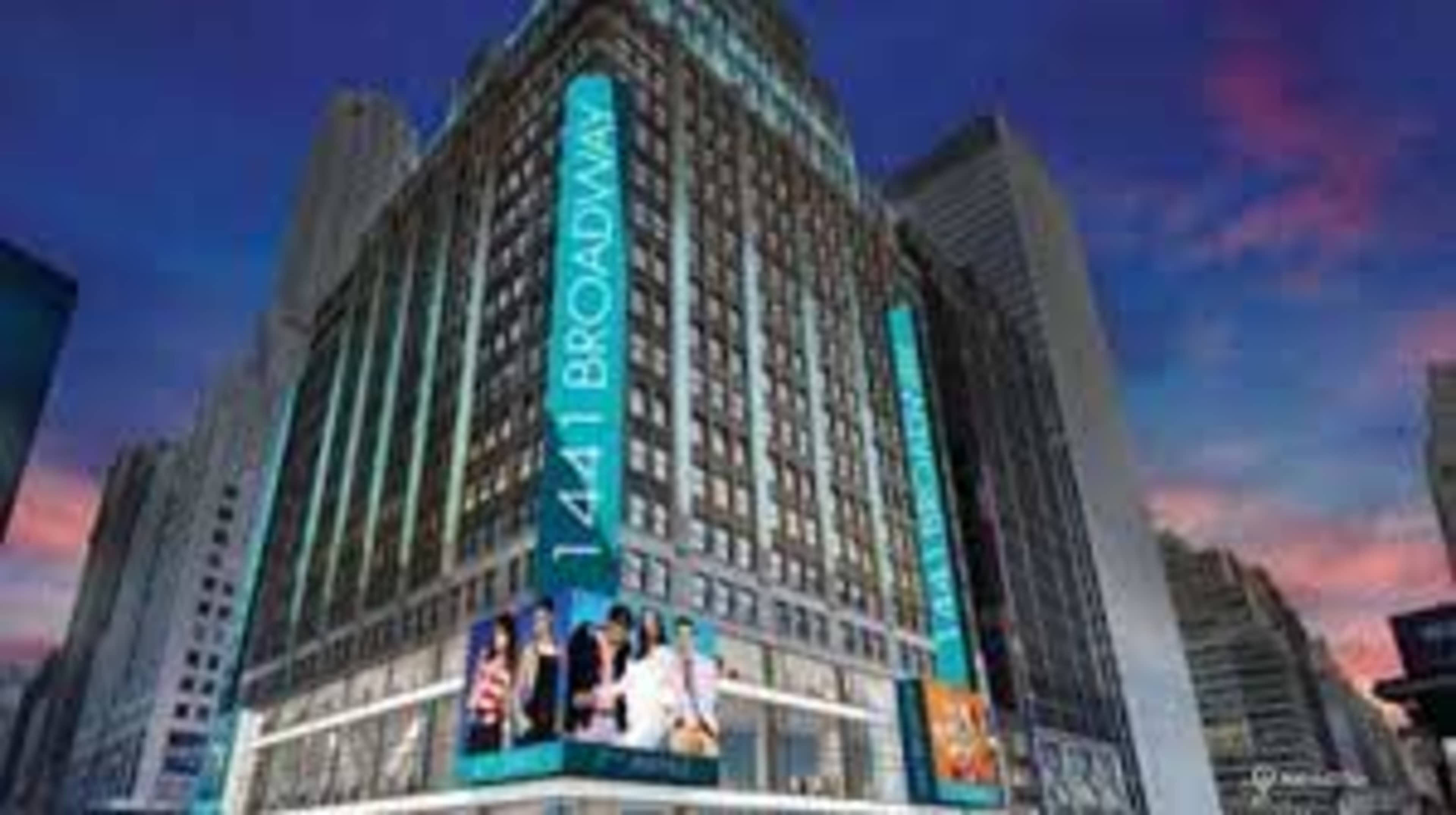 A large, illuminated building at 1441 Broadway, surrounded by other skyscrapers and a colorful sky at dusk.