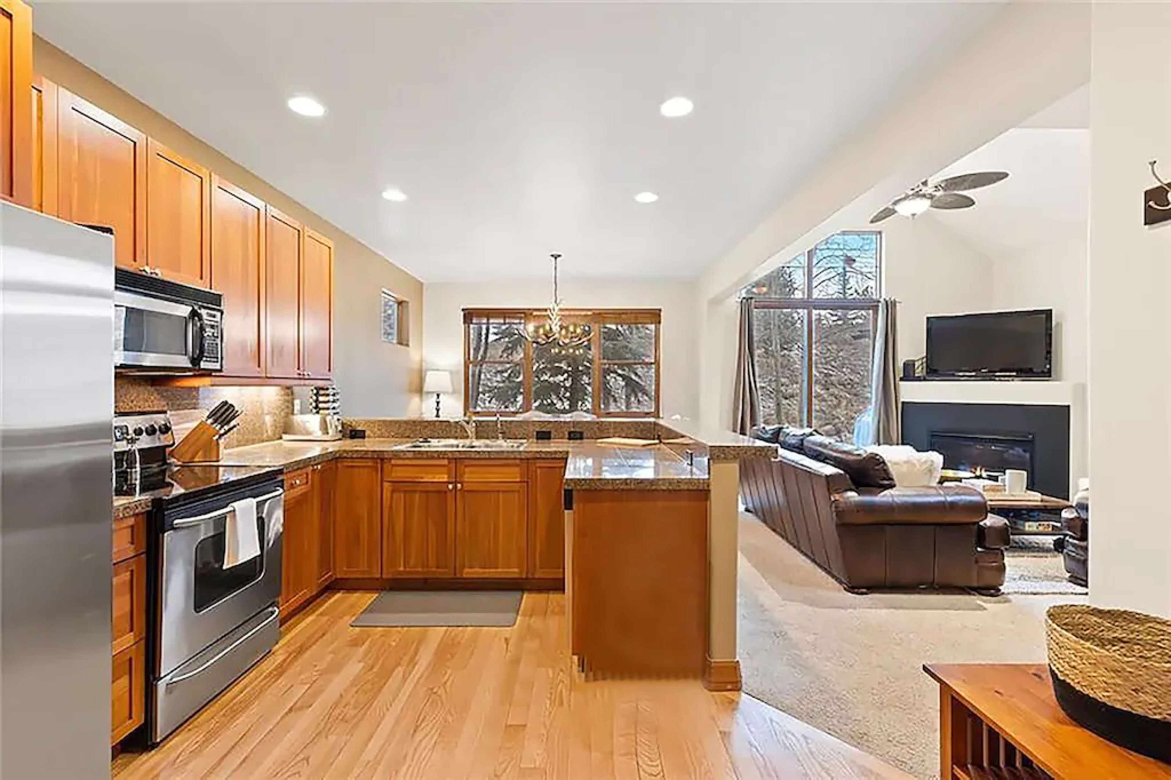 A modern kitchen features wooden cabinets, granite countertops, and stainless steel appliances, connected to a living area with large windows and a fireplace.