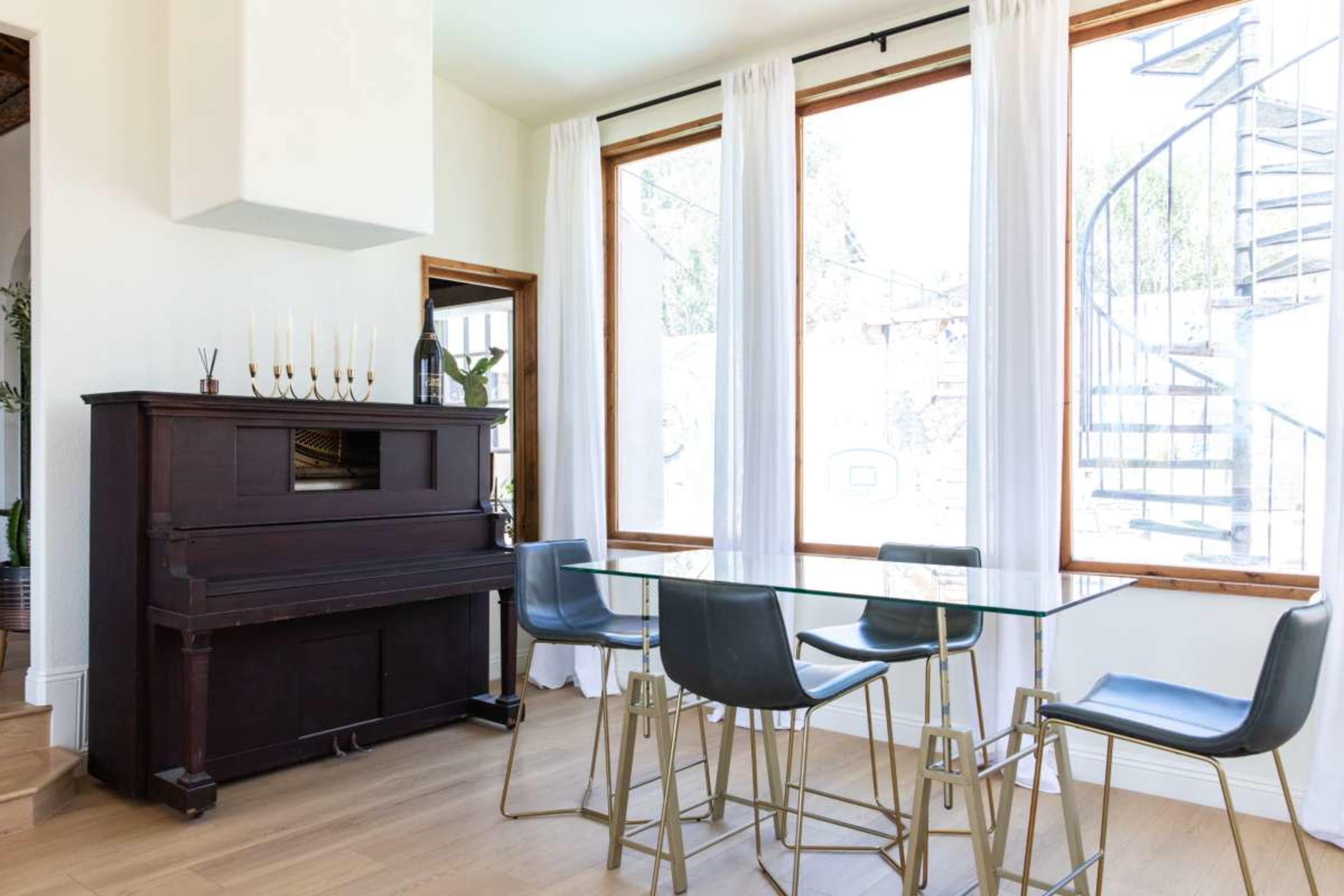 The image shows a dining area with a glass table and four metal chairs, alongside a dark wooden piano and large windows with natural light.