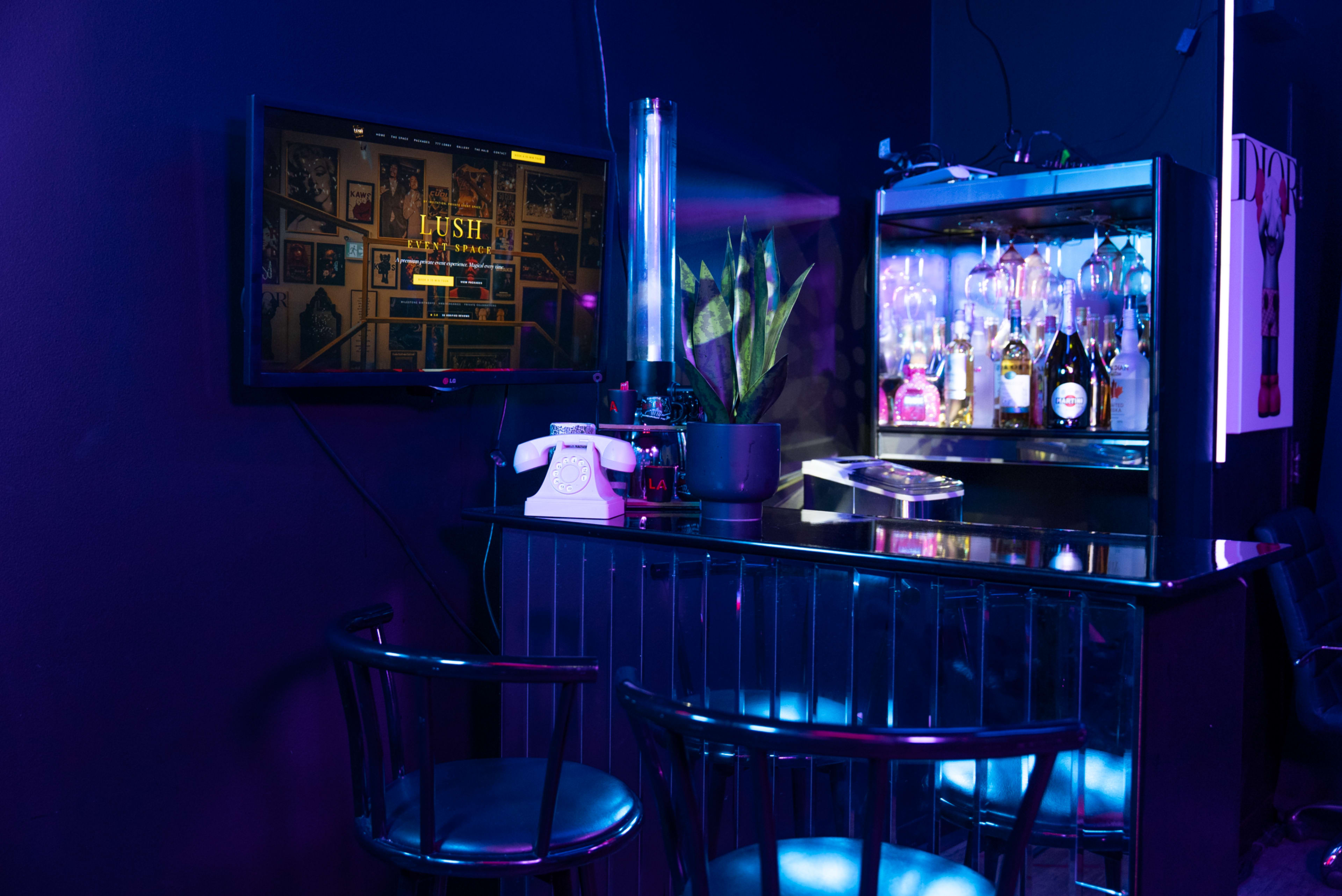 A dimly lit bar area features a modern shelf displaying various bottles, a touchscreen monitor mounted on the wall, and two black chairs positioned around a sleek counter.