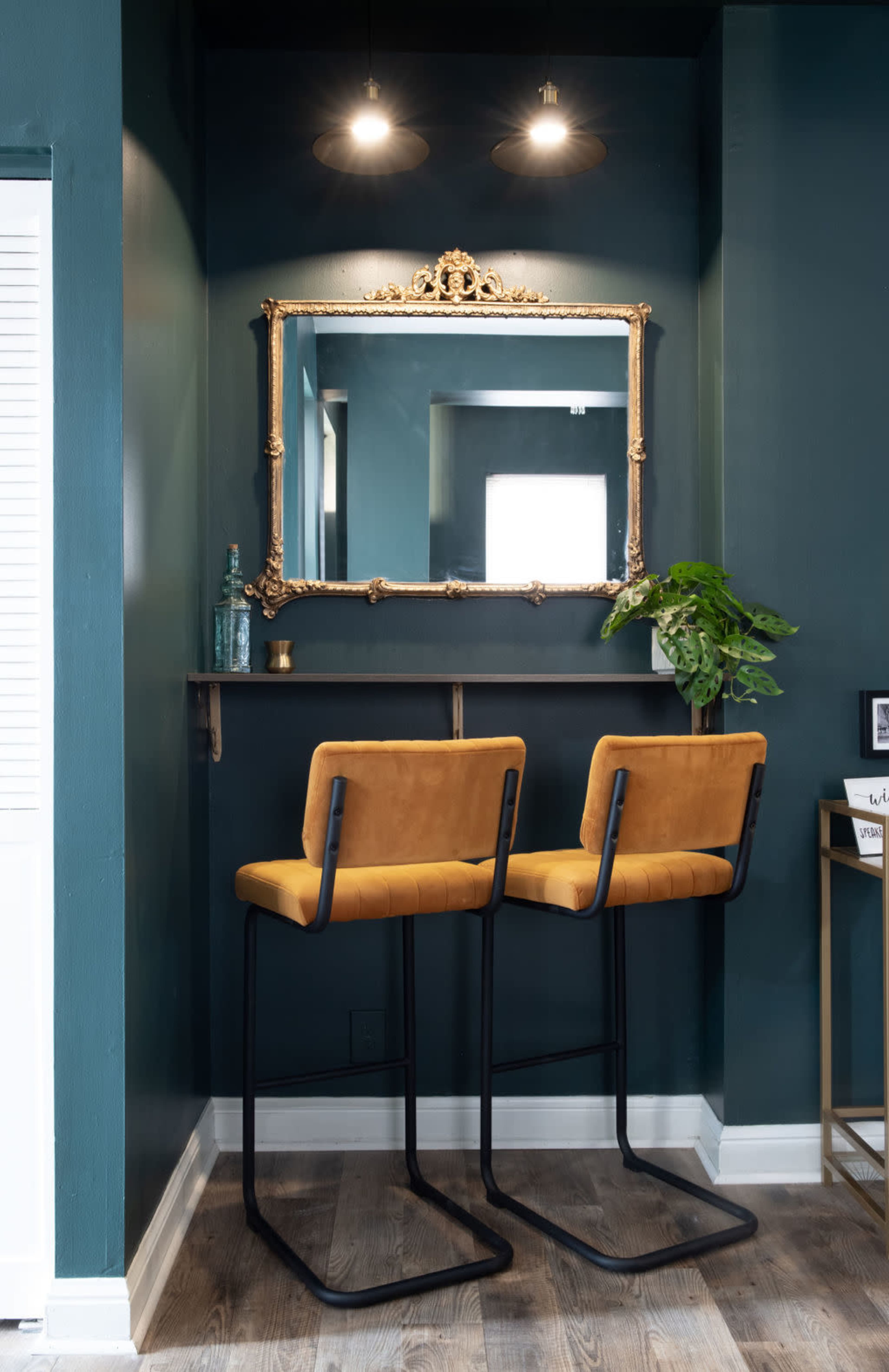 A well-lit area features two mustard-colored bar stools positioned at a wall-mounted shelf with a large ornate mirror above it, against a dark green wall.