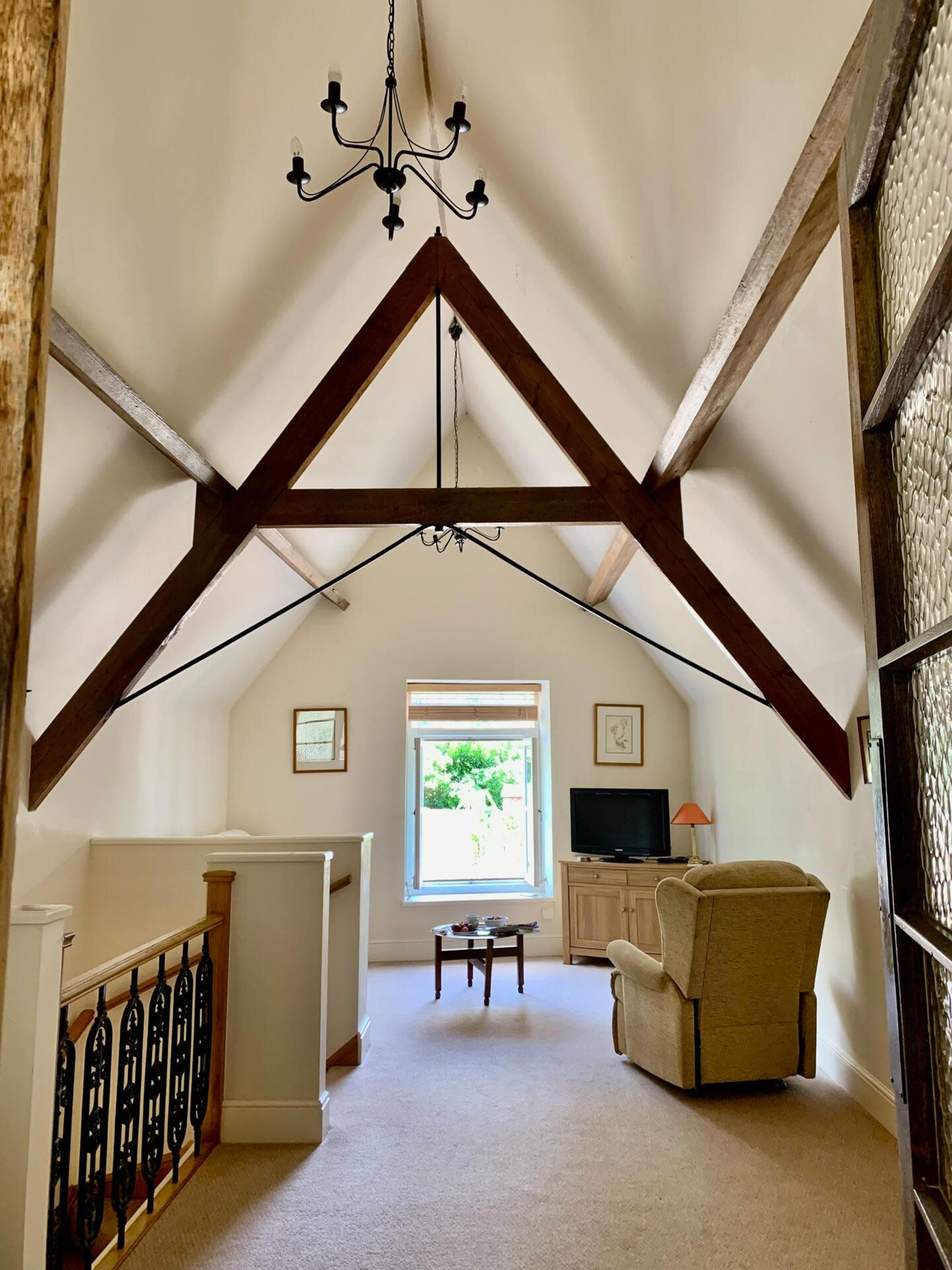 A spacious interior with a vaulted ceiling featuring wooden beams, a small seating area with a chair, and a window overlooking greenery.