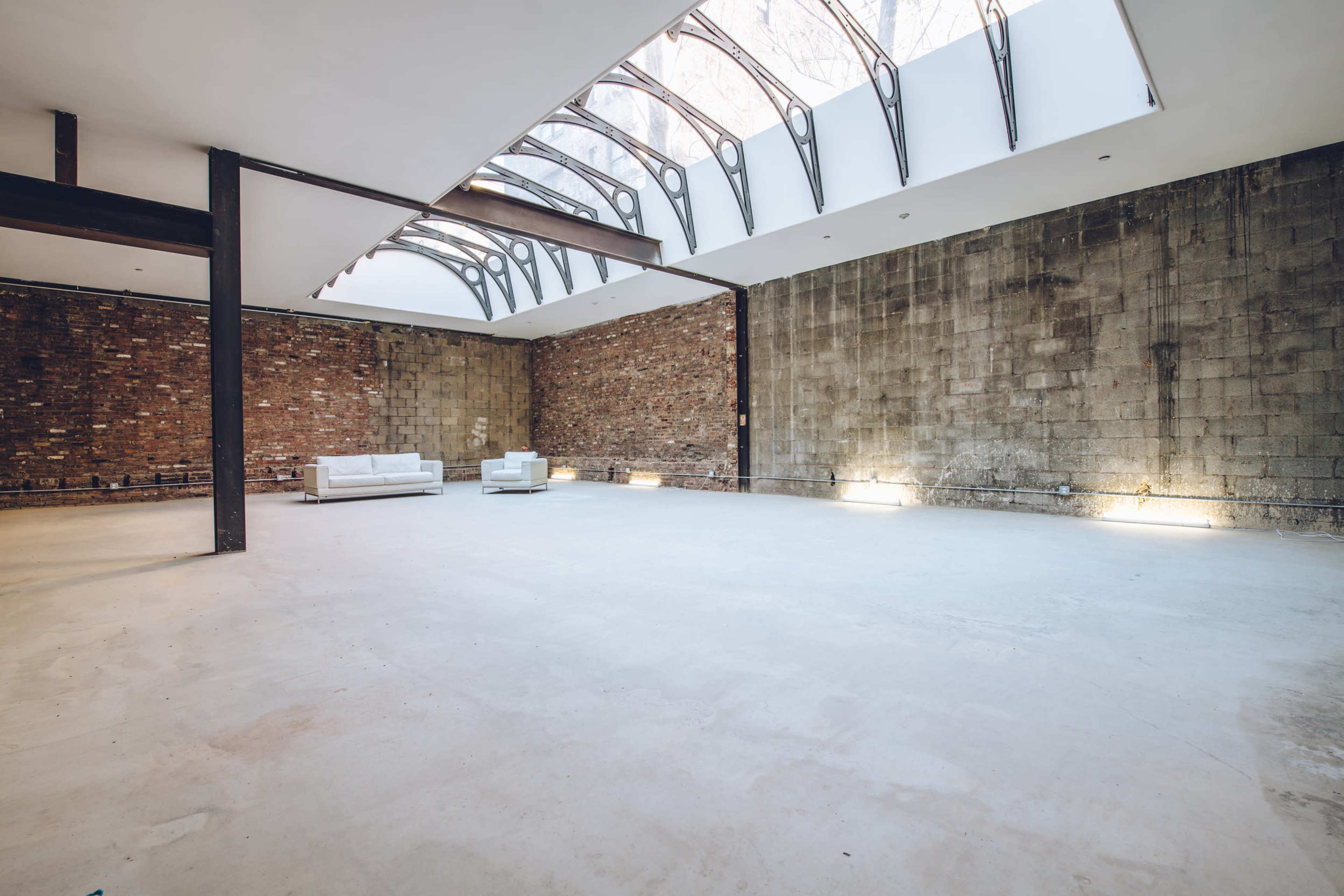 The image shows a spacious interior with exposed brick walls, large windows, and minimal furnishings, featuring two white sofas.